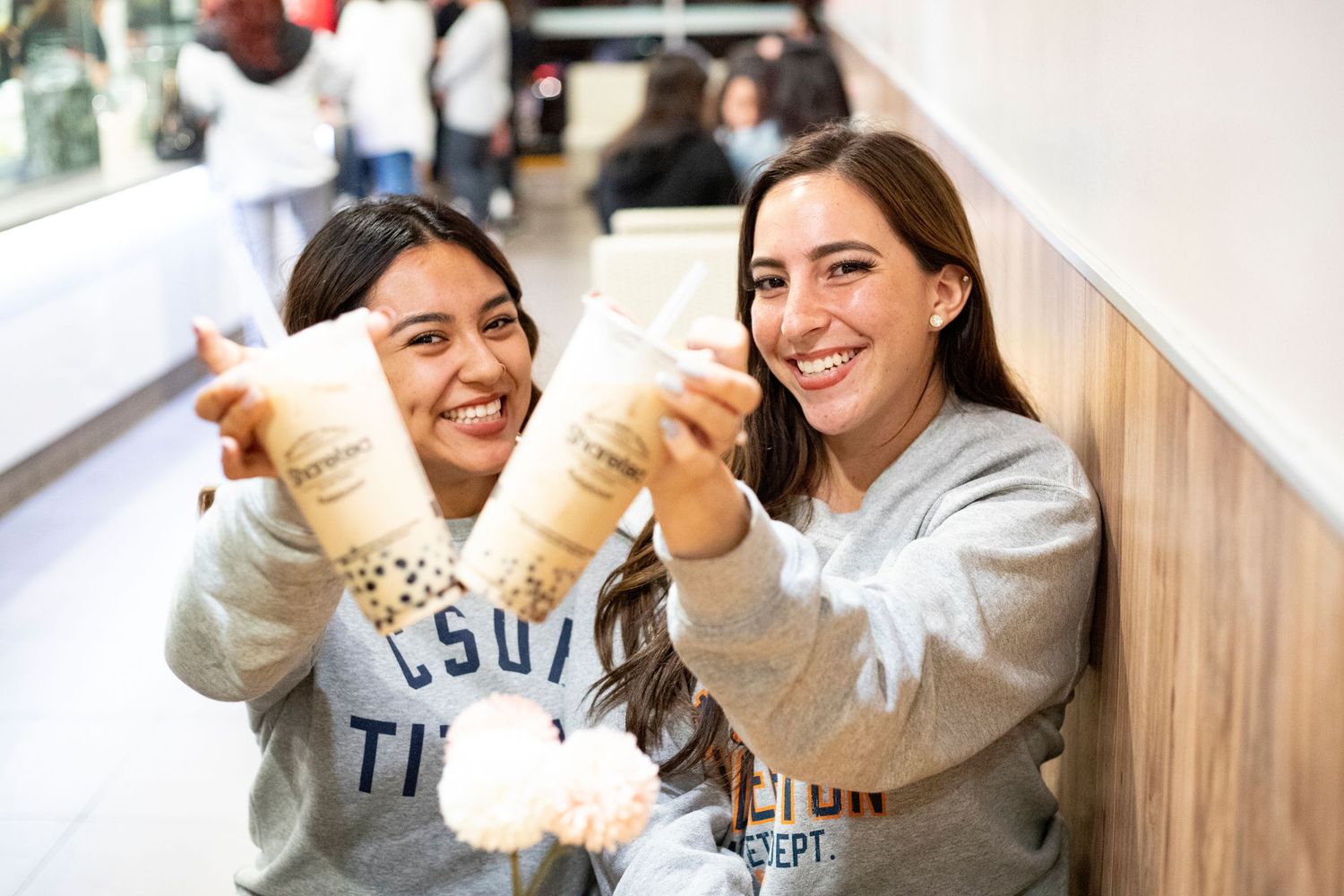 Two young friends are sitting at at table at Sharetea. They are smiling at the camera as they hold their boba tea for the camera
