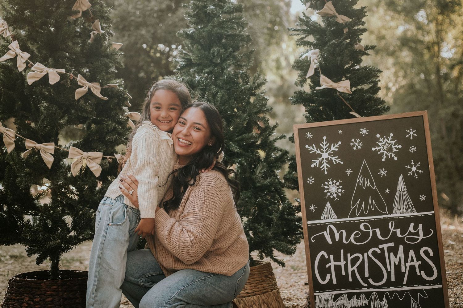 Mother and daughter smiling together at a Christmas tree farm with a Merry Christmas chalkboard sign.