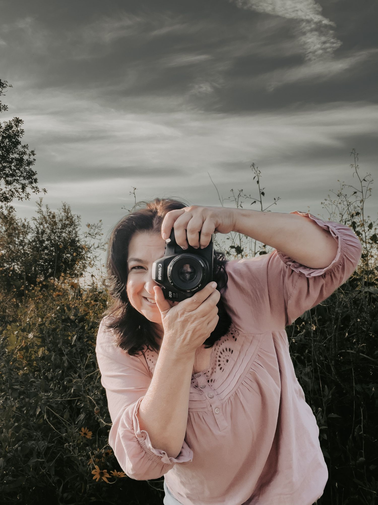photographer in a pink shirt is holding a DSLR camera and smiling as she points it at the camera to take a picture