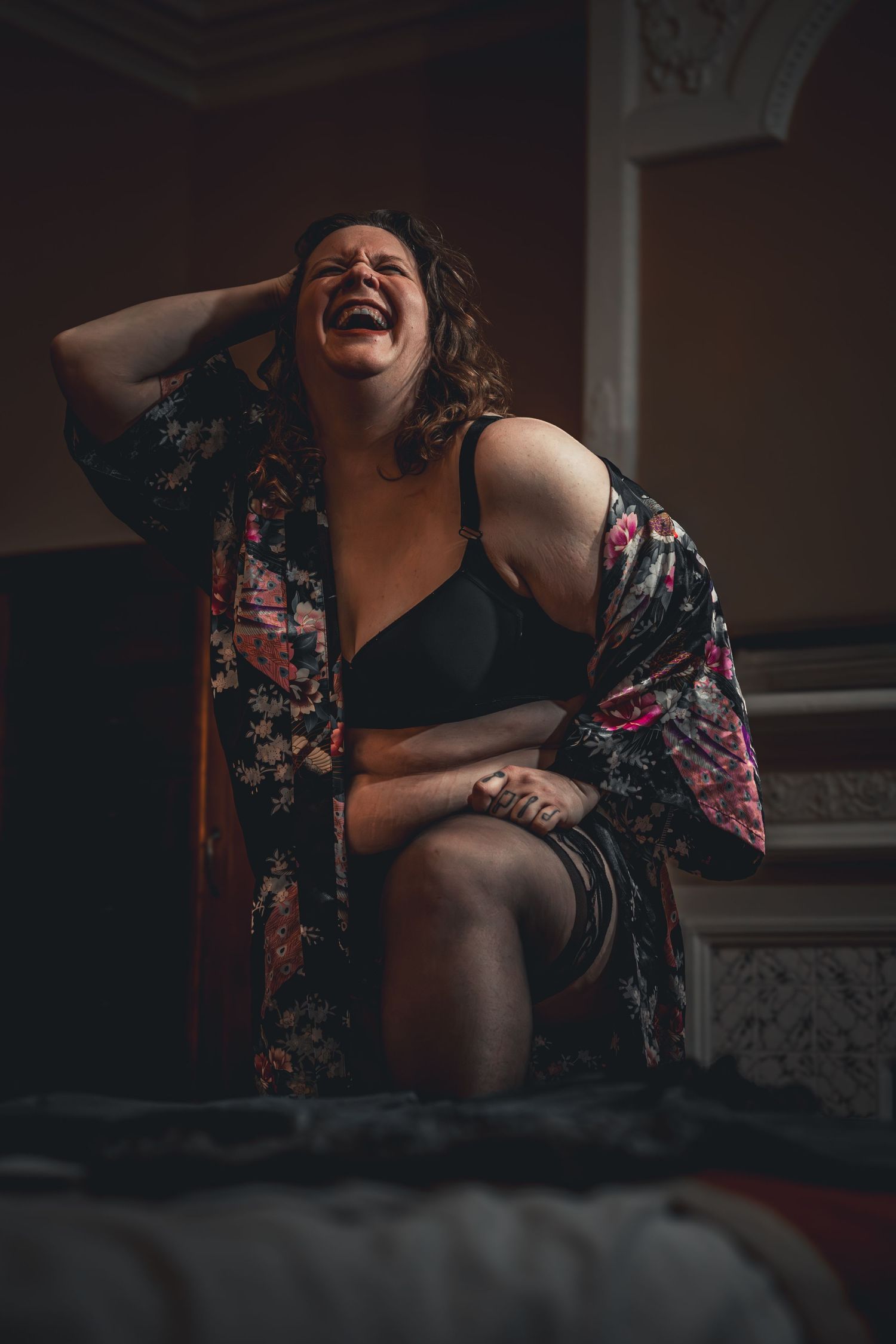 Woman in floral robe and black bra laughing joyfully while sitting on a bed in a dimly lit room.