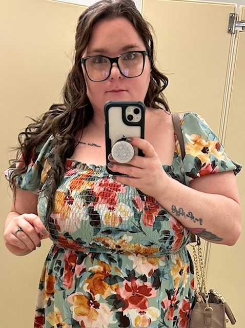 A changing room selfie showing a woman with long brown curly hair wearing colorful floral dress taking a photo in the mirror with her cell.