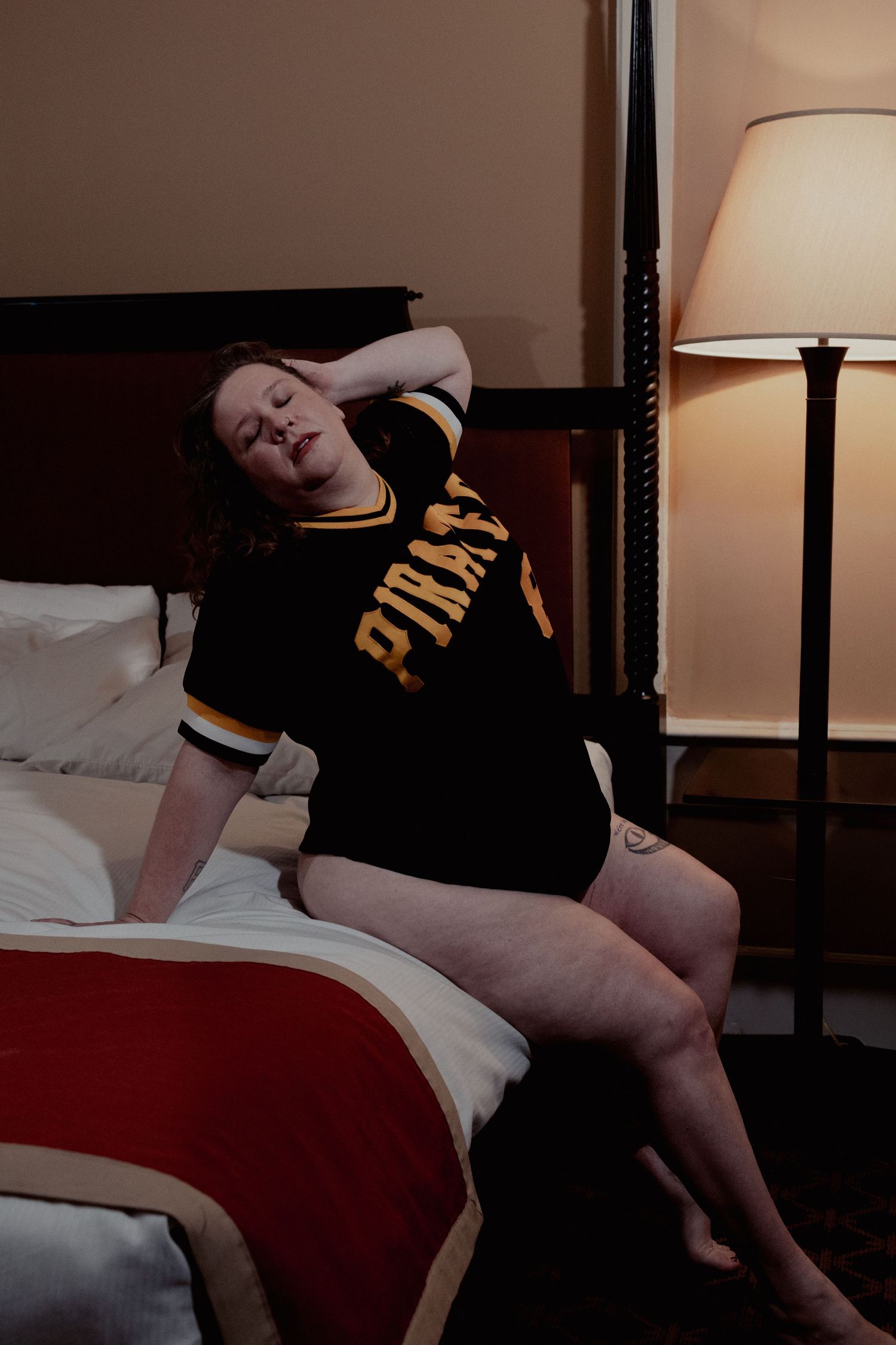 A person in a black shirt lies asleep on a bed with white sheets and a red blanket in a dimly lit room.