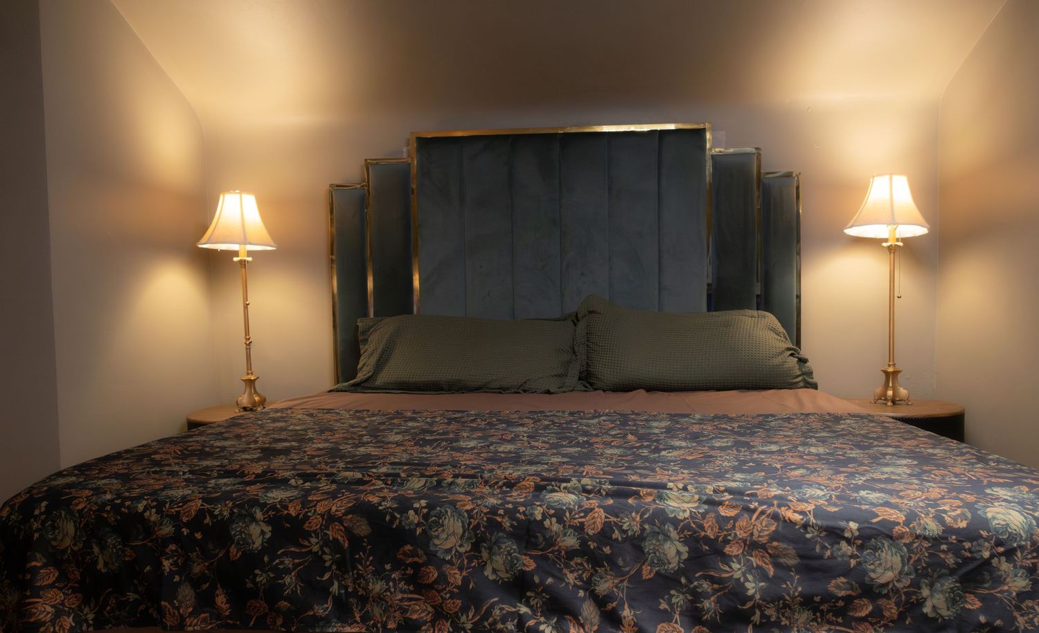 Elegant hotel bedroom with floral bedspread, blue velvet headboard, and warm brass table lamps on both sides.