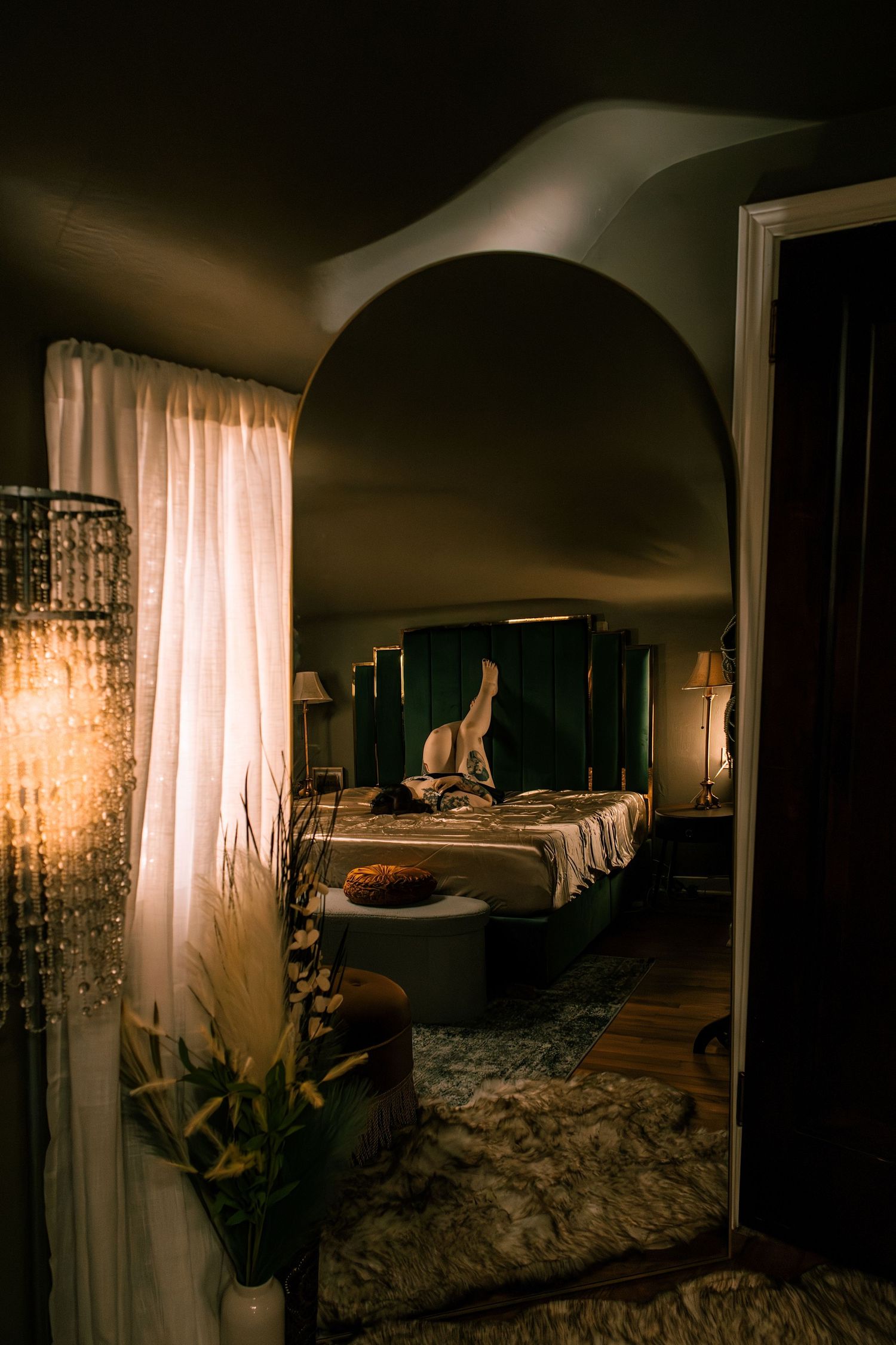Dimly lit bedroom with arched doorway, green velvet headboard, sheer curtains, and warm ambient lighting.