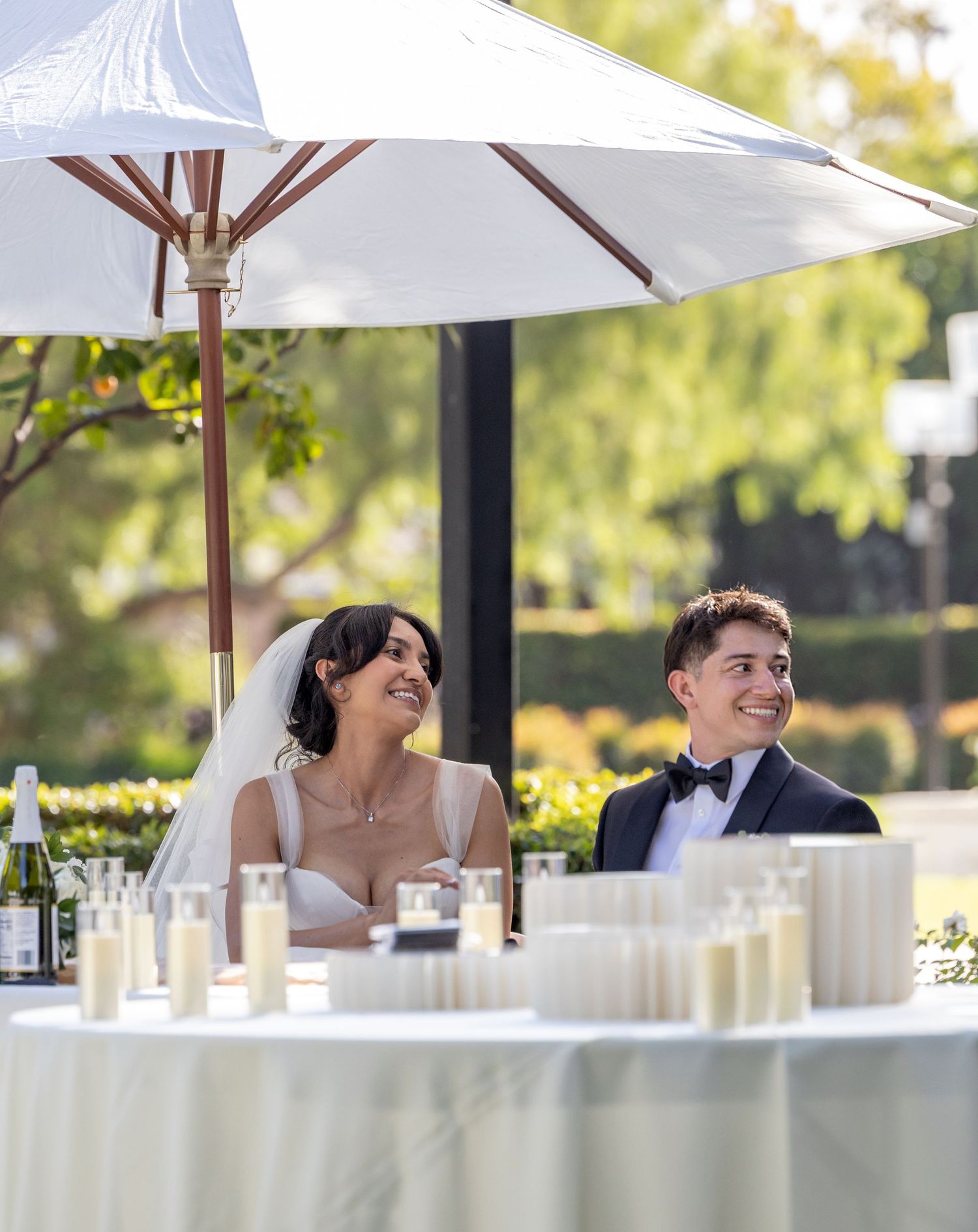 Wedding couple seated under white umbrella at outdoor reception table.