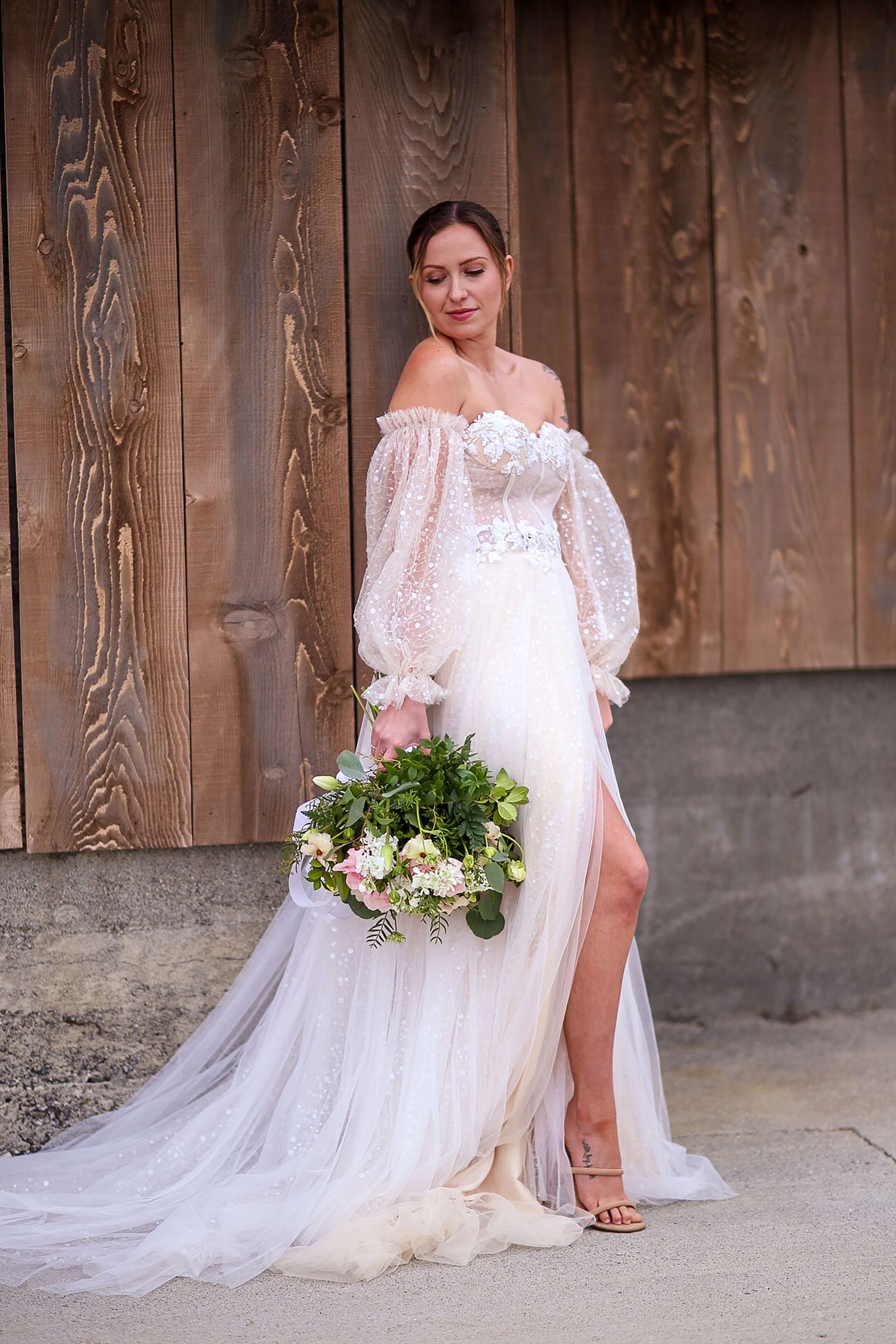 Bride in lace gown with flowing train sitting beside rustic wooden wall.