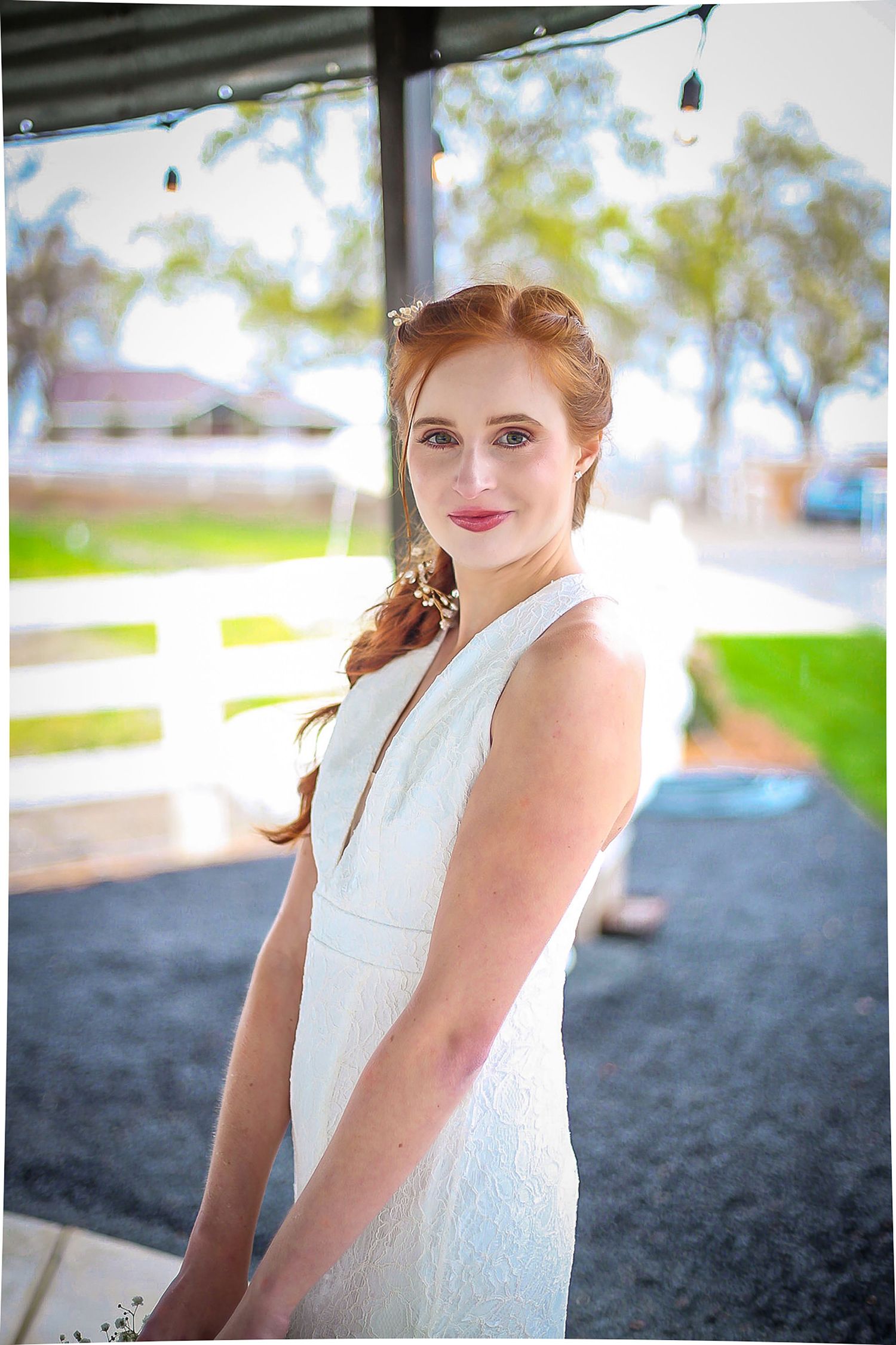 Bride in elegant white dress standing under covered porch on sunny day.