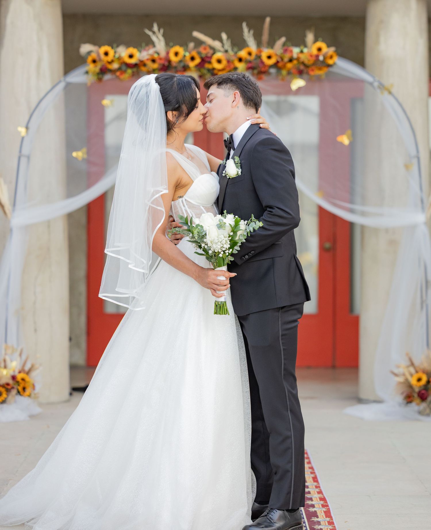 A couple shares a romantic kiss during their outdoor wedding ceremony decorated with sunflowers and white drapes.