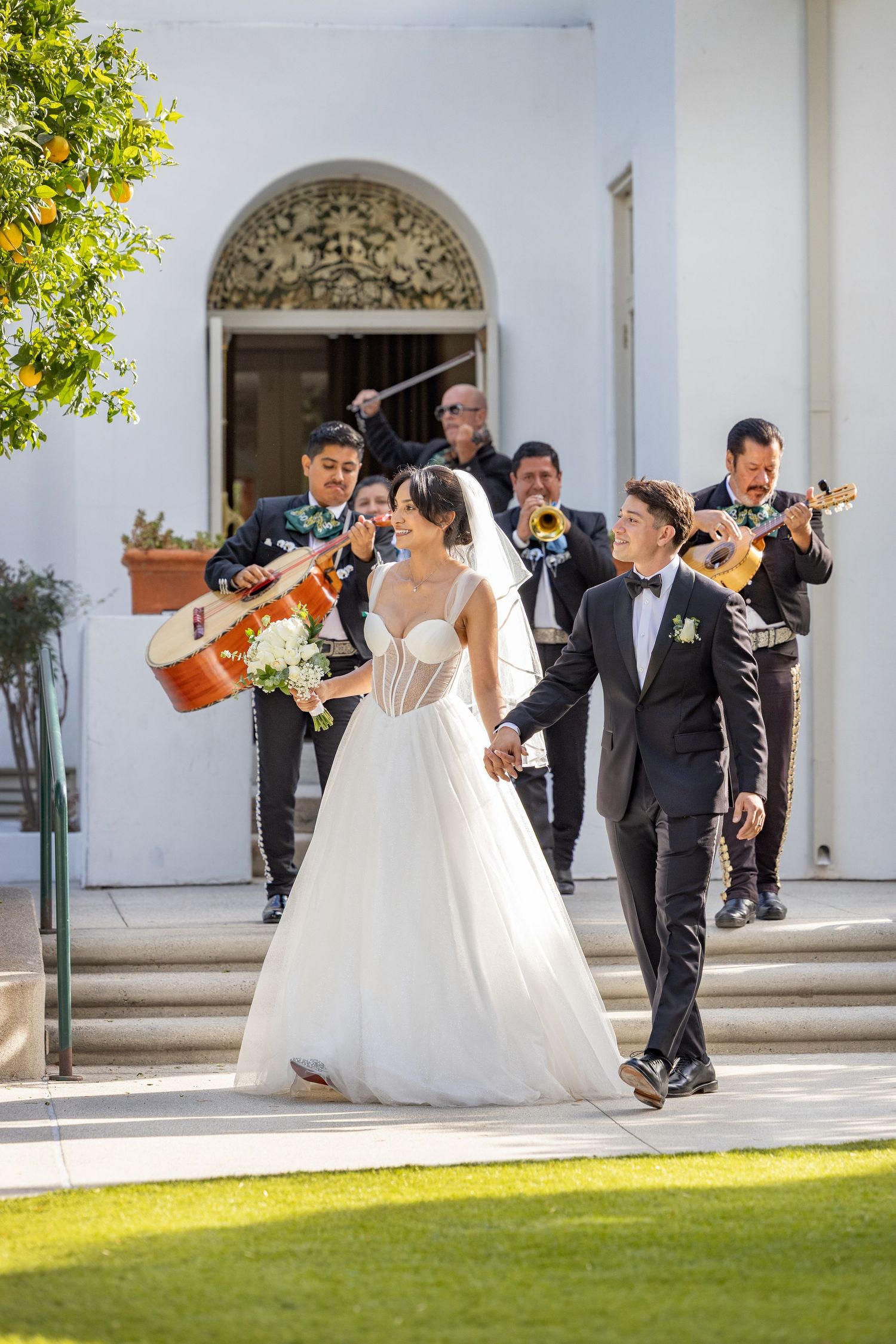 Wedding party members in formal attire strike playful poses against white arches and green lawn at outdoor venue.