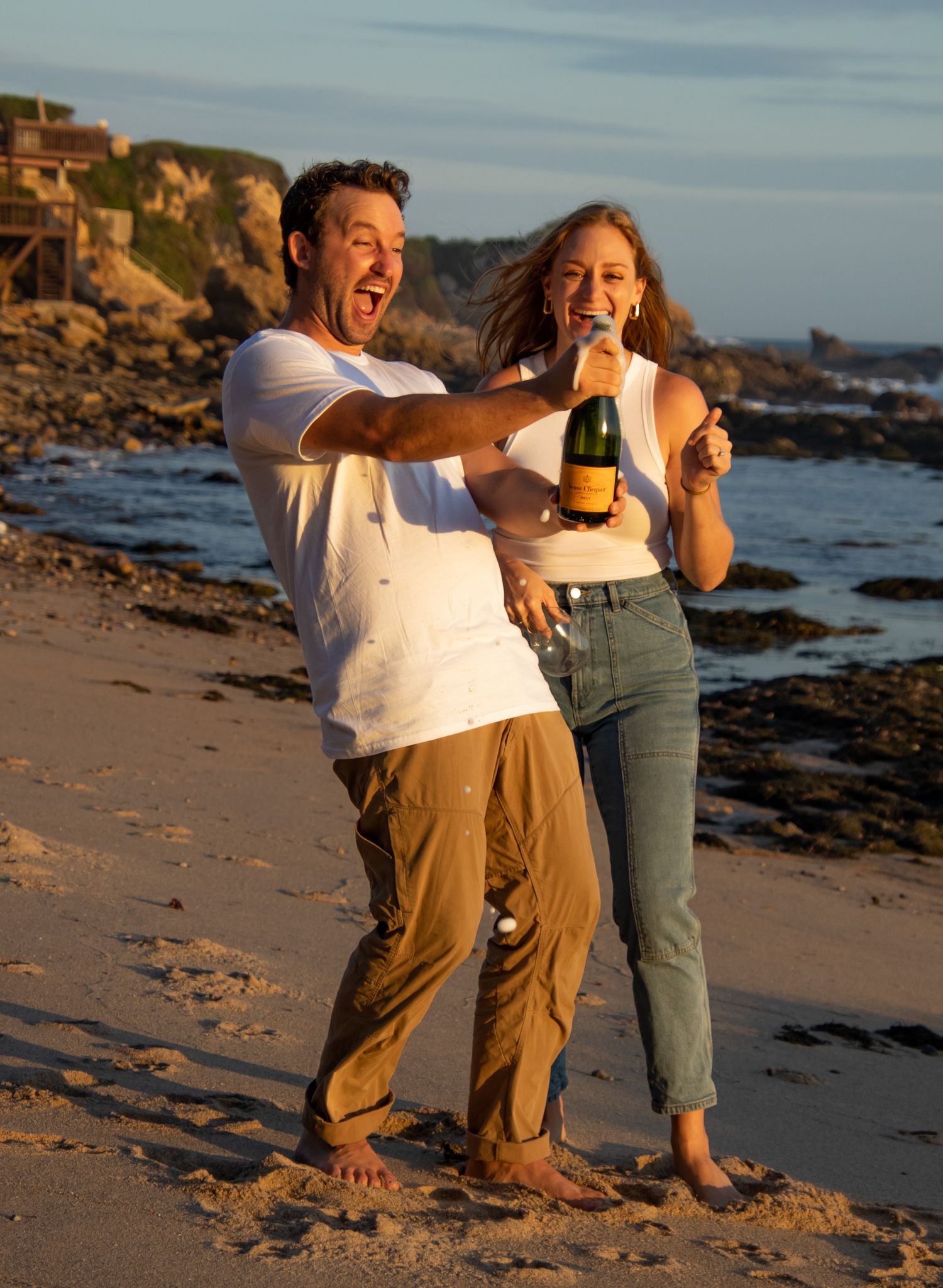 A couple laughs together while enjoying a sunset on a rocky beach with waves crashing in the background.