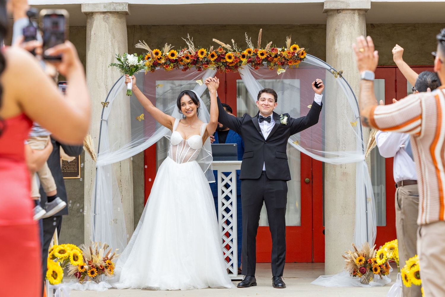A wedding party celebrates with arms raised under decorative floral arrangements in an outdoor venue.