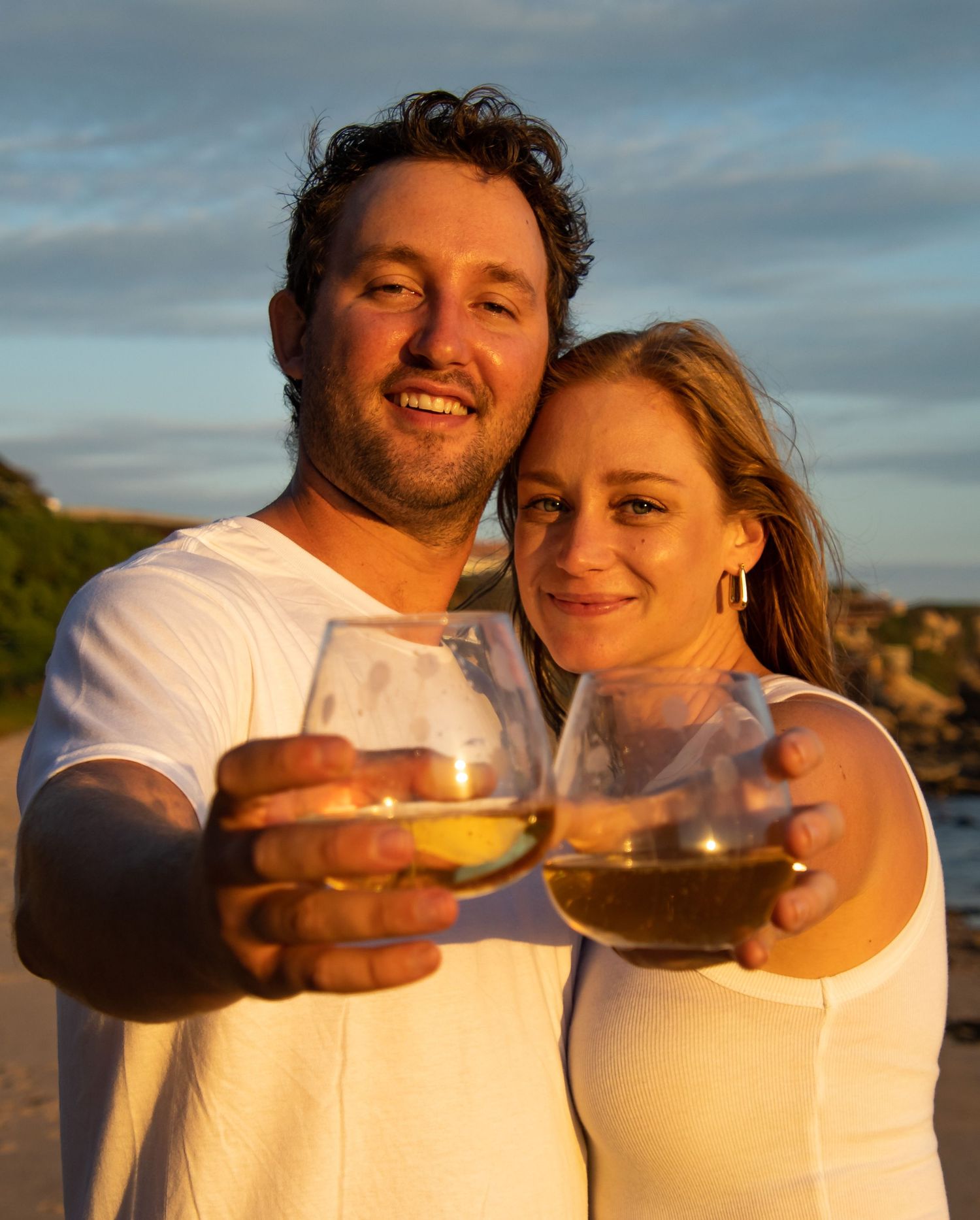 A couple toasts with wine glasses during golden hour outdoors.