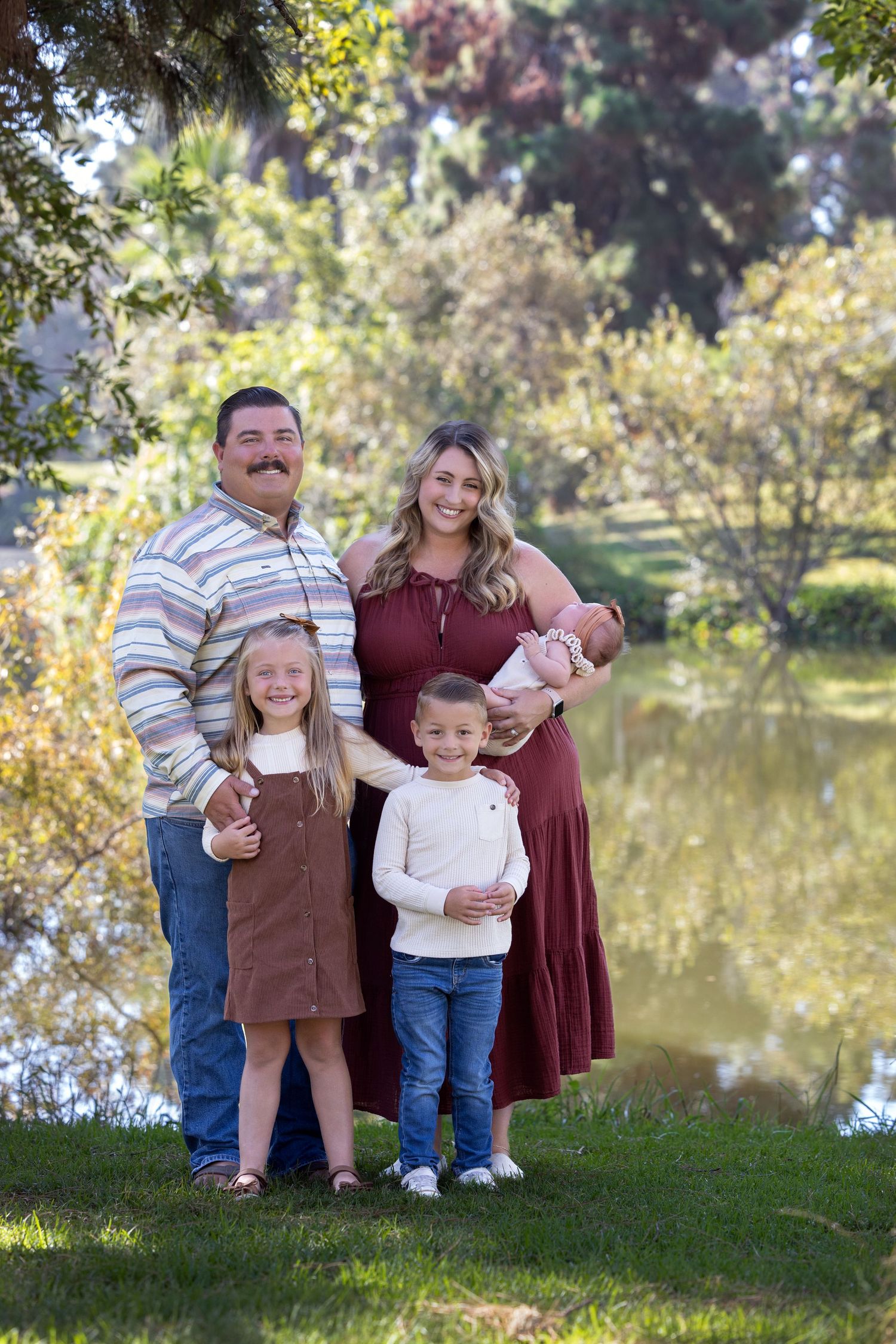 A family poses together on a grassy hill by a pond during a sunny autumn day for outdoor portraits.