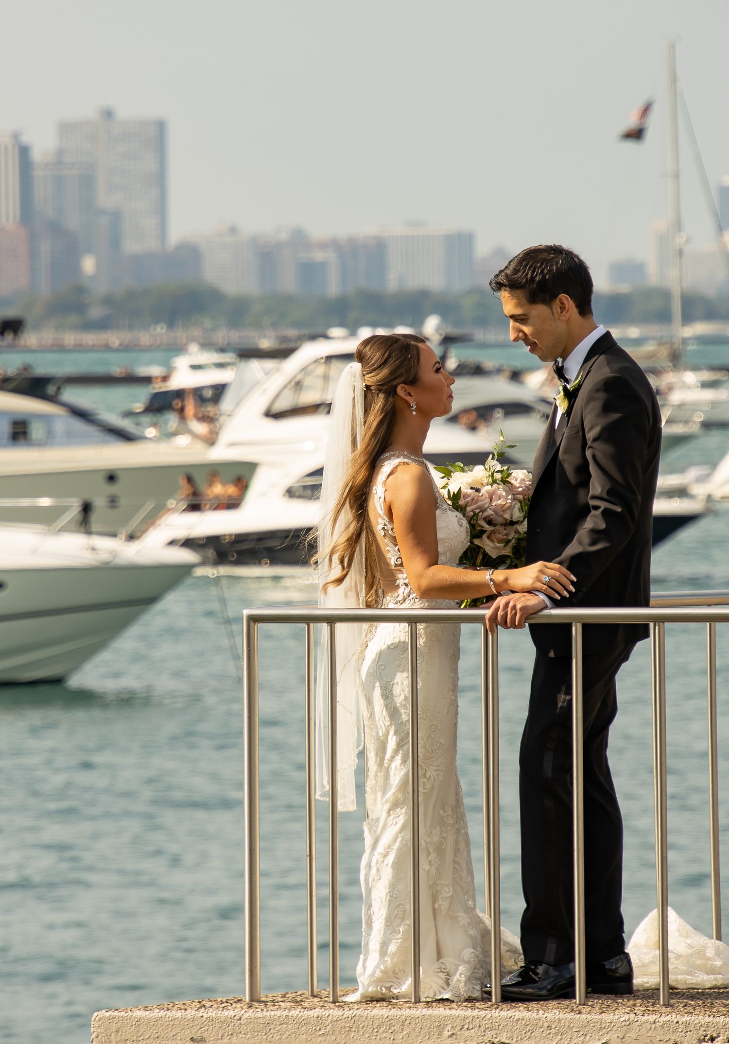 A couple in wedding attire share a romantic moment at a marina with boats and city skyline in the background.