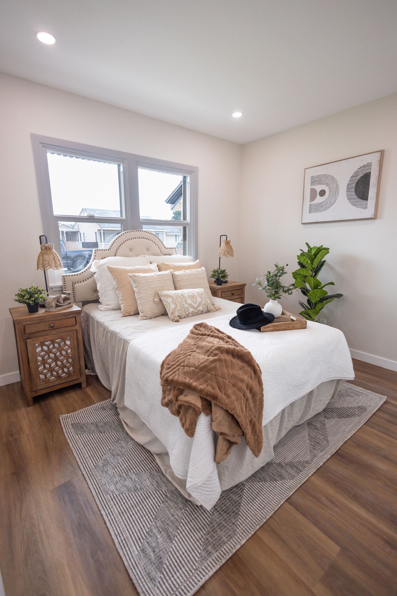 Modern bedroom with white bedding, brown throw blanket, decorative pillows, and wooden nightstand on hardwood floors.