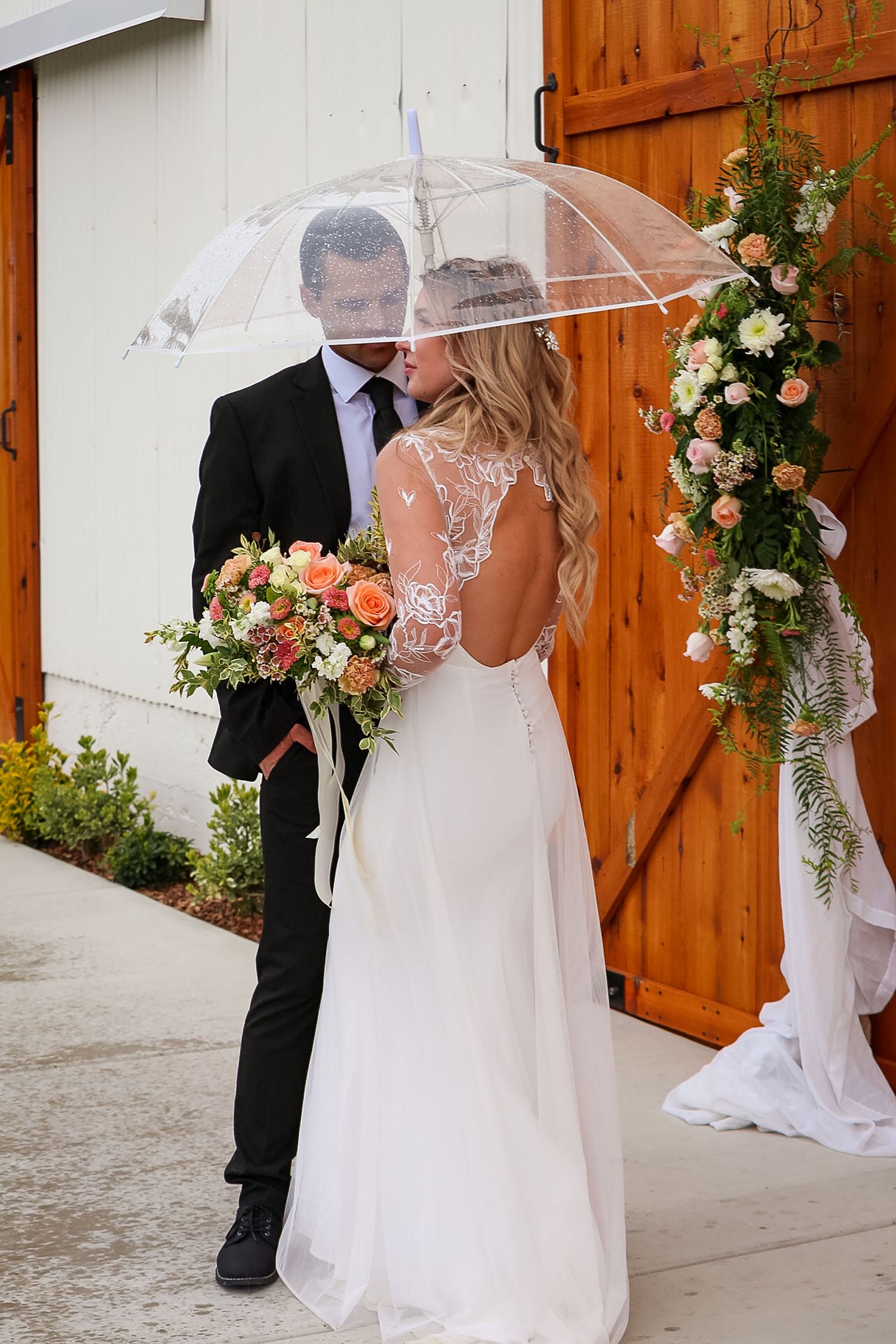 Couple sharing a clear umbrella in front of wooden doors with floral decorations at a wedding ceremony.