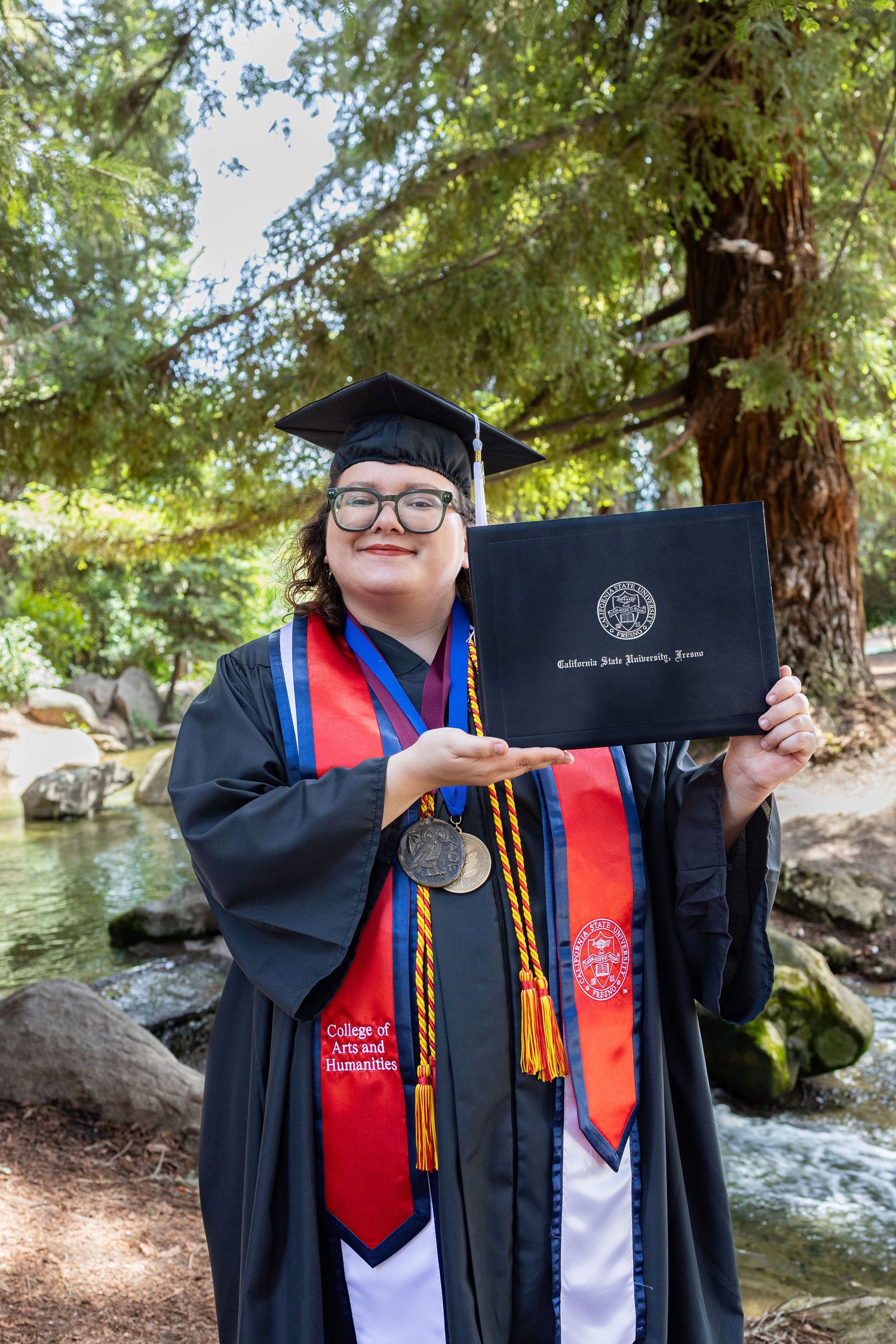 A graduate in cap and gown poses with their diploma and honor cords against a scenic outdoor backdrop with trees and water.