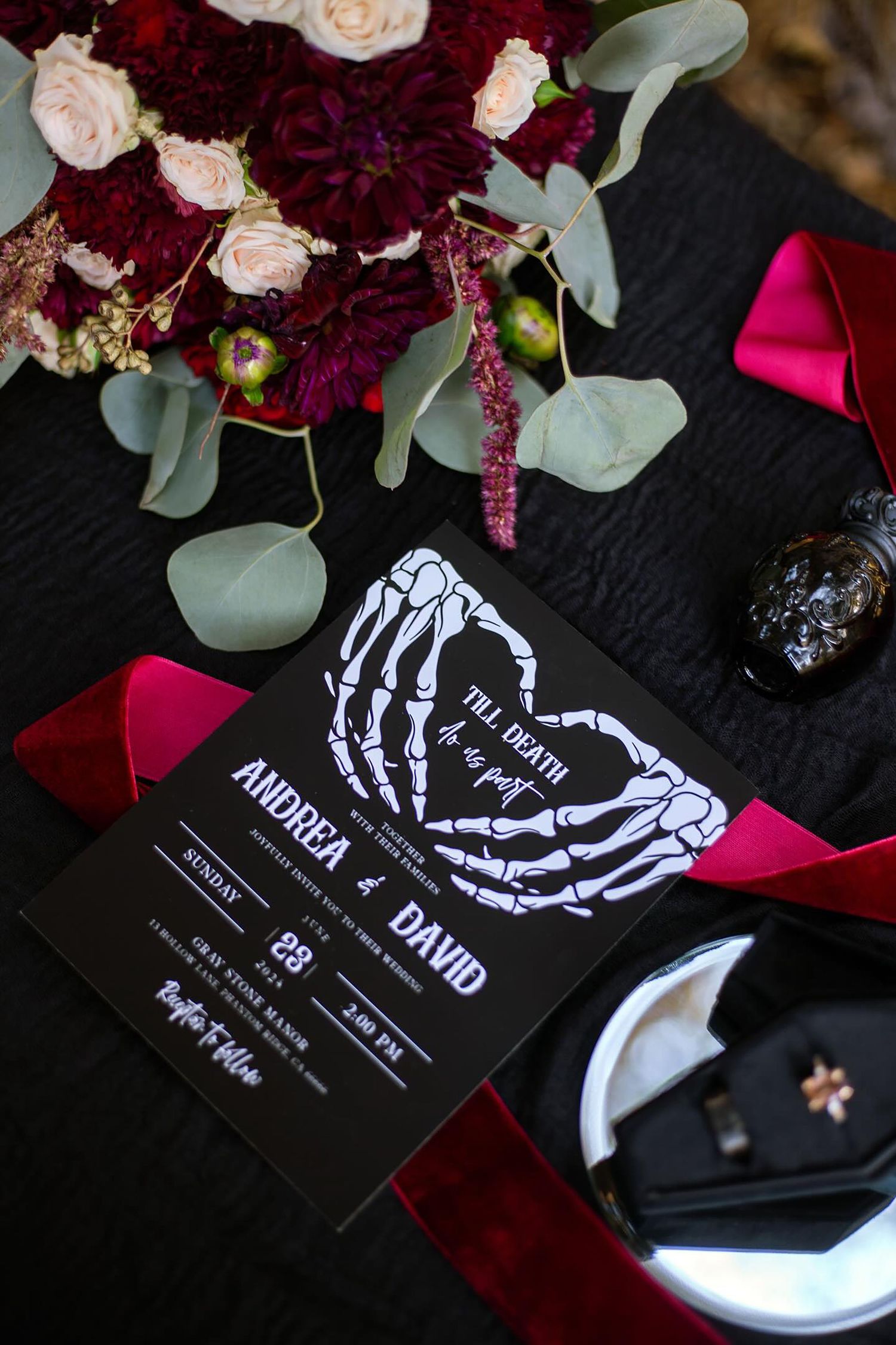 Gothic wedding invitations with skeleton hands design displayed among burgundy flowers.