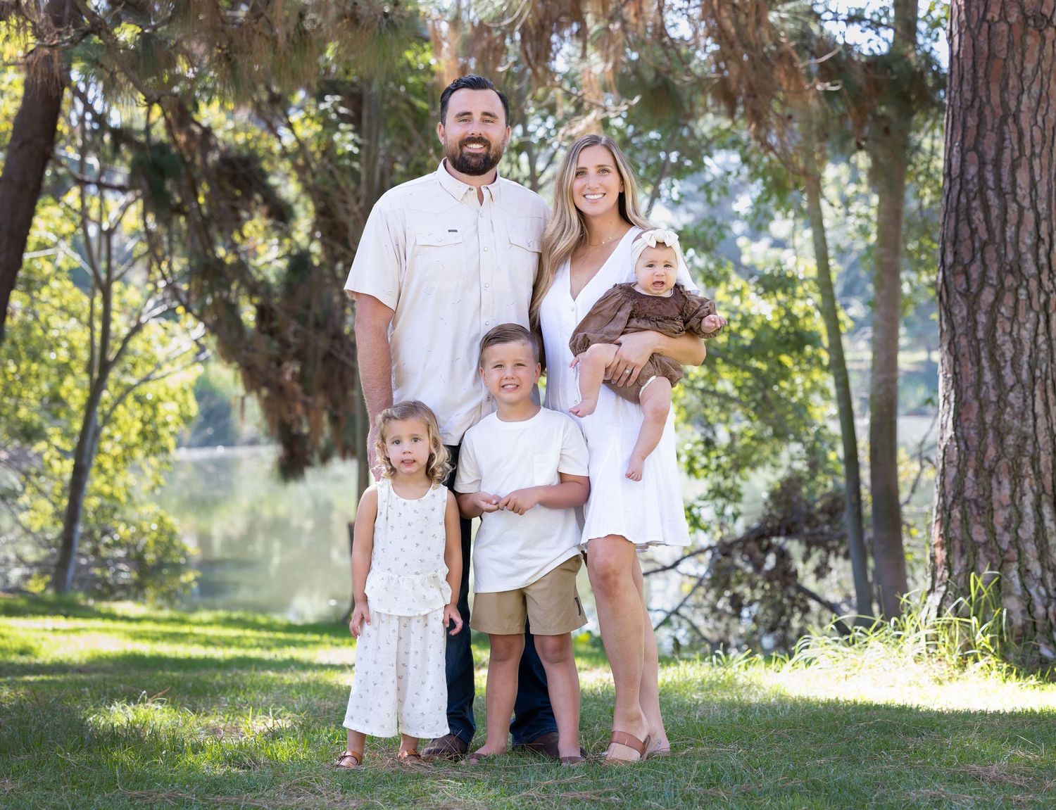 A family of five poses together outdoors in a park setting with trees and sunlight filtering through the branches.