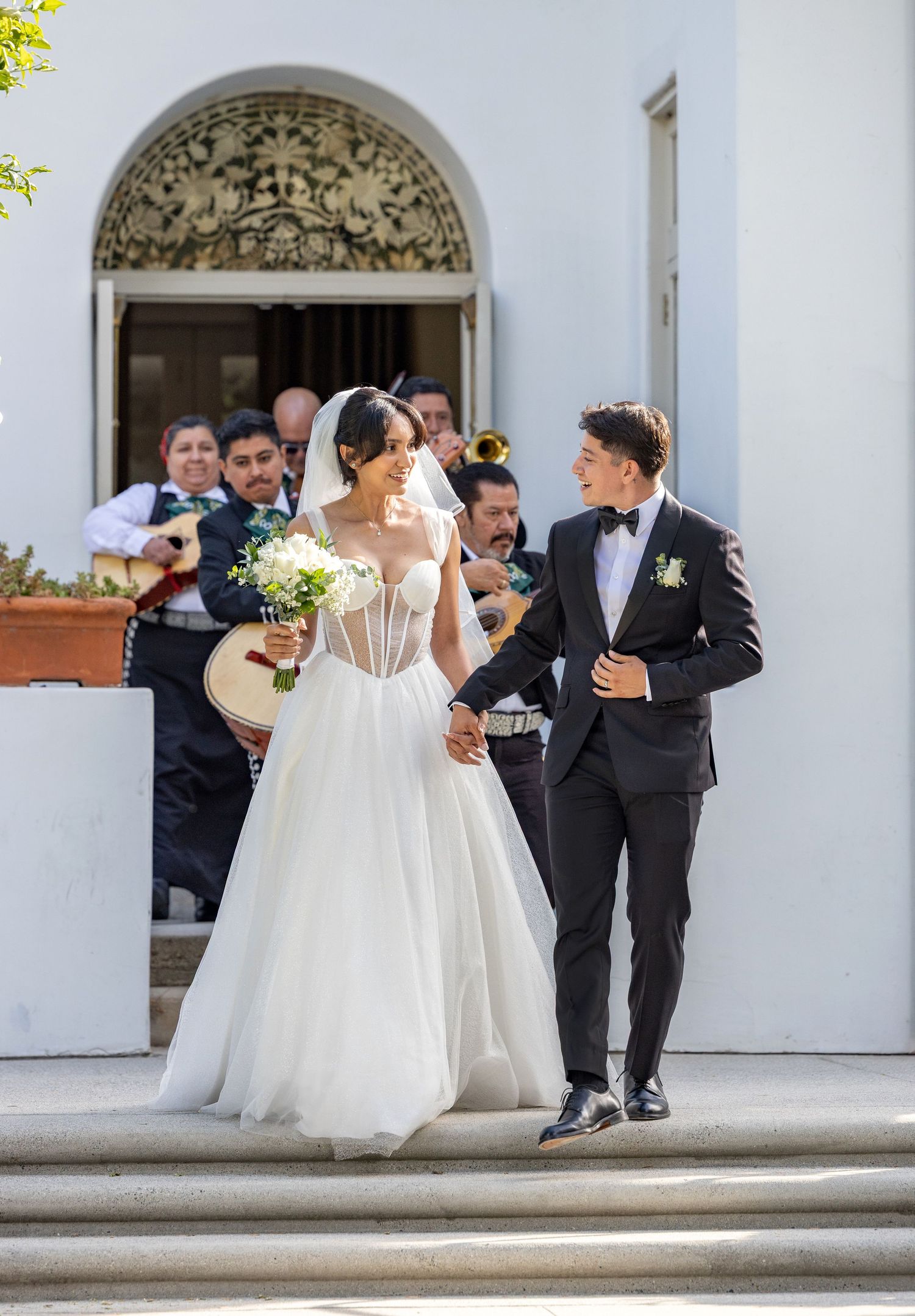 A newlywed couple exits a church while mariachi musicians perform in the background on a sunny day.