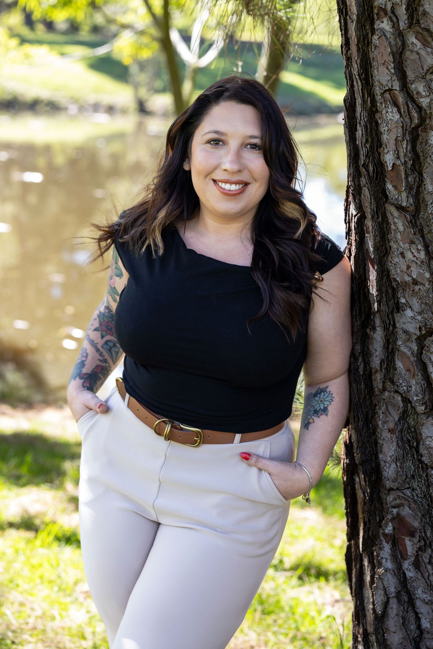 A person in a black top and white skirt poses outdoors near a tree trunk with a pond in the background.