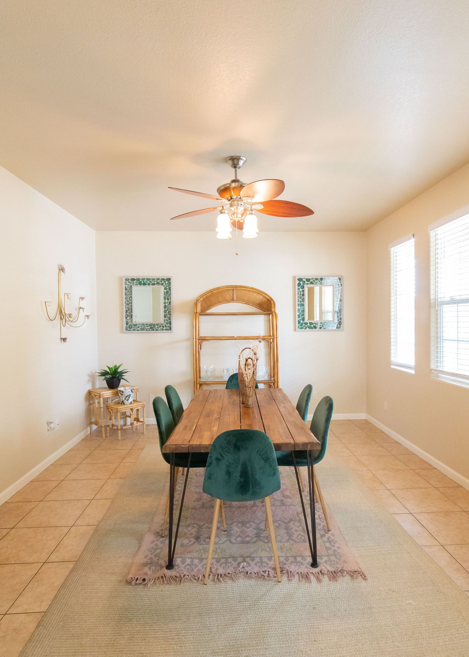 Modern dining room with wooden table, emerald velvet chairs, ceiling fan, and beige tile flooring in an open layout space.