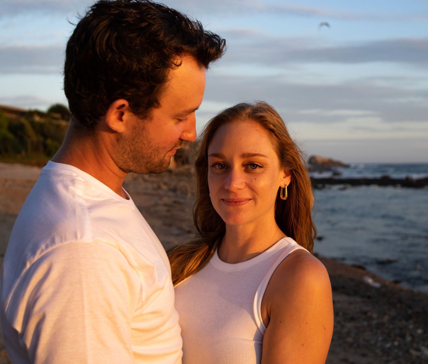 A couple shares an intimate moment at sunset on a rocky beach with ocean waves in the background.