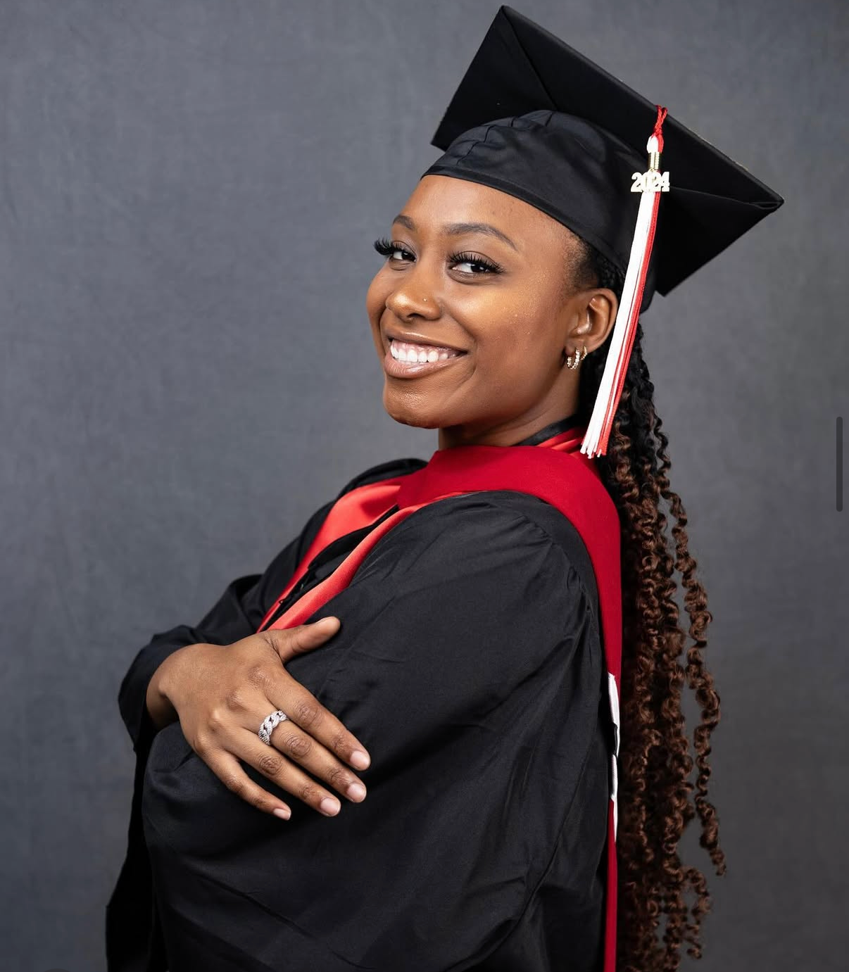 A graduate in cap and gown smiles while posing for a portrait against a gray background.