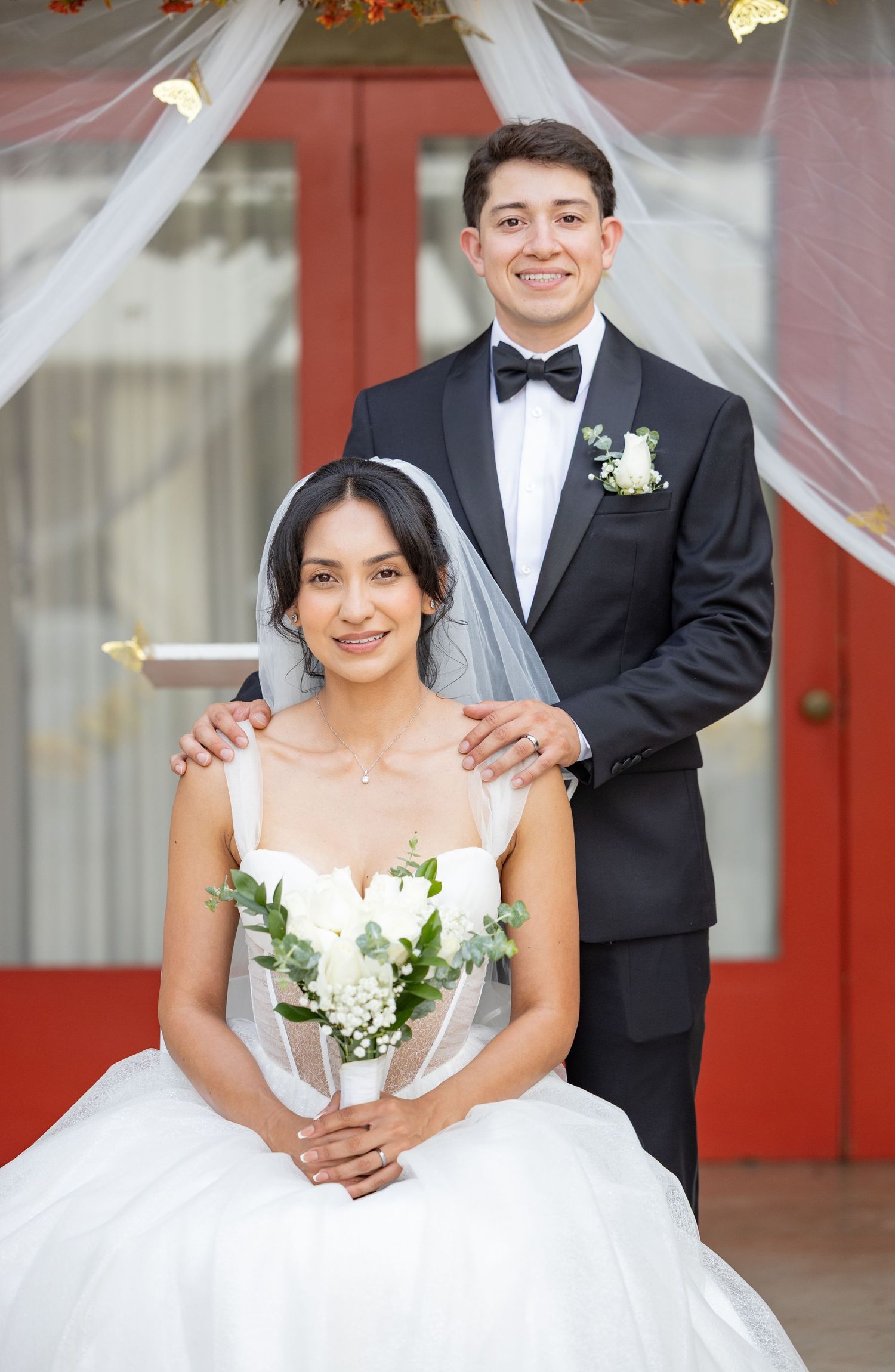 Wedding couple poses in front of red doors wearing traditional formal attire with white bouquet.