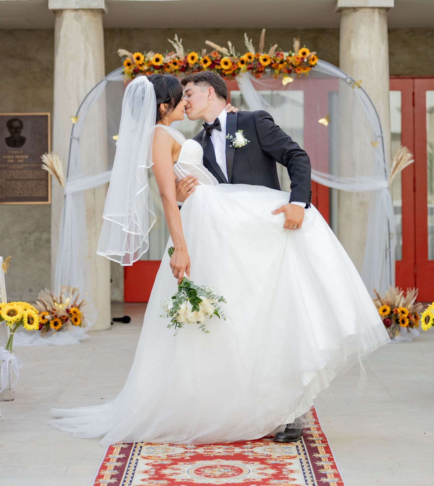 A romantic wedding scene with a couple kissing under an arch decorated with sunflowers and white tulle fabric.