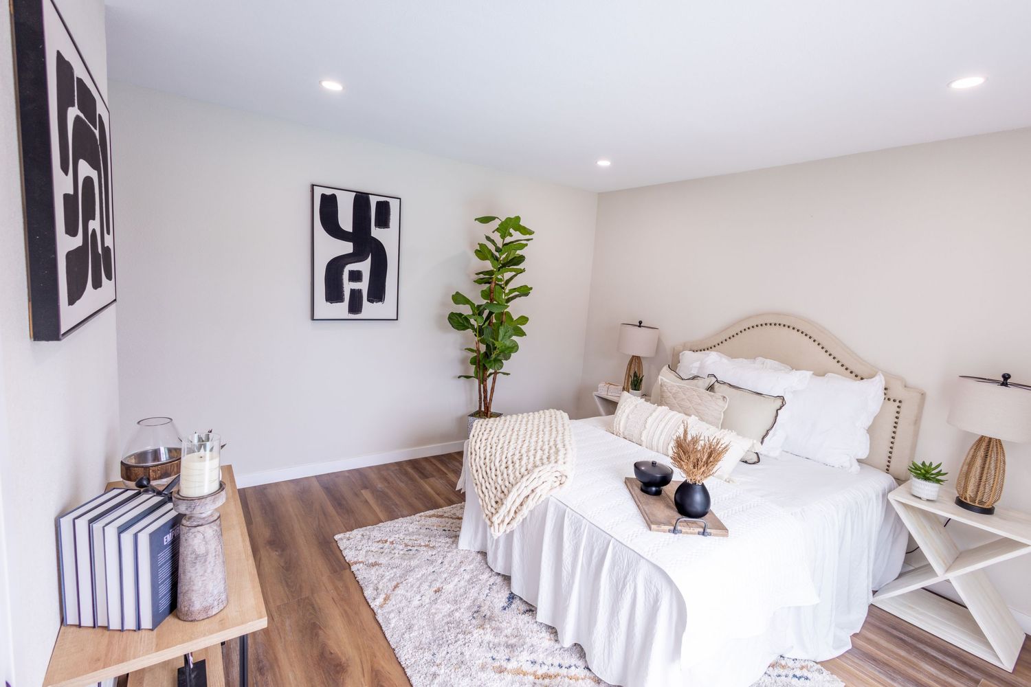 A modern white bedroom with black and white artwork, natural wood accents, and decorative plants in the corner.