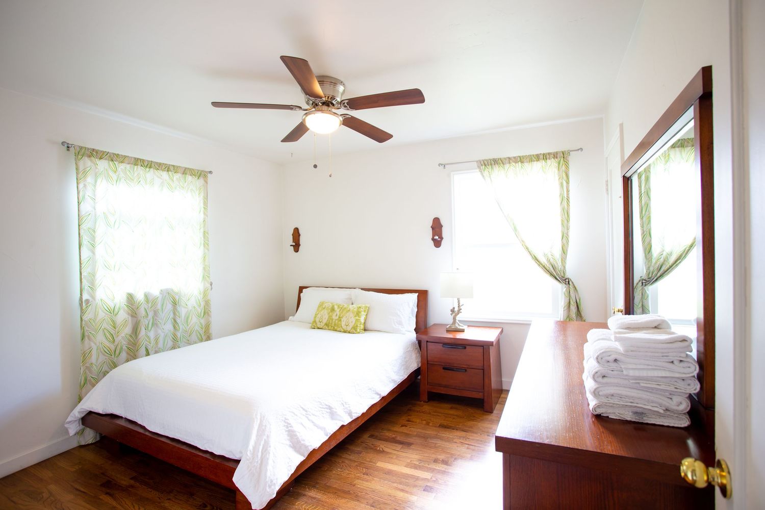 Bright bedroom interior with hardwood floors, white walls, ceiling fan, and sheer curtains letting in natural light.