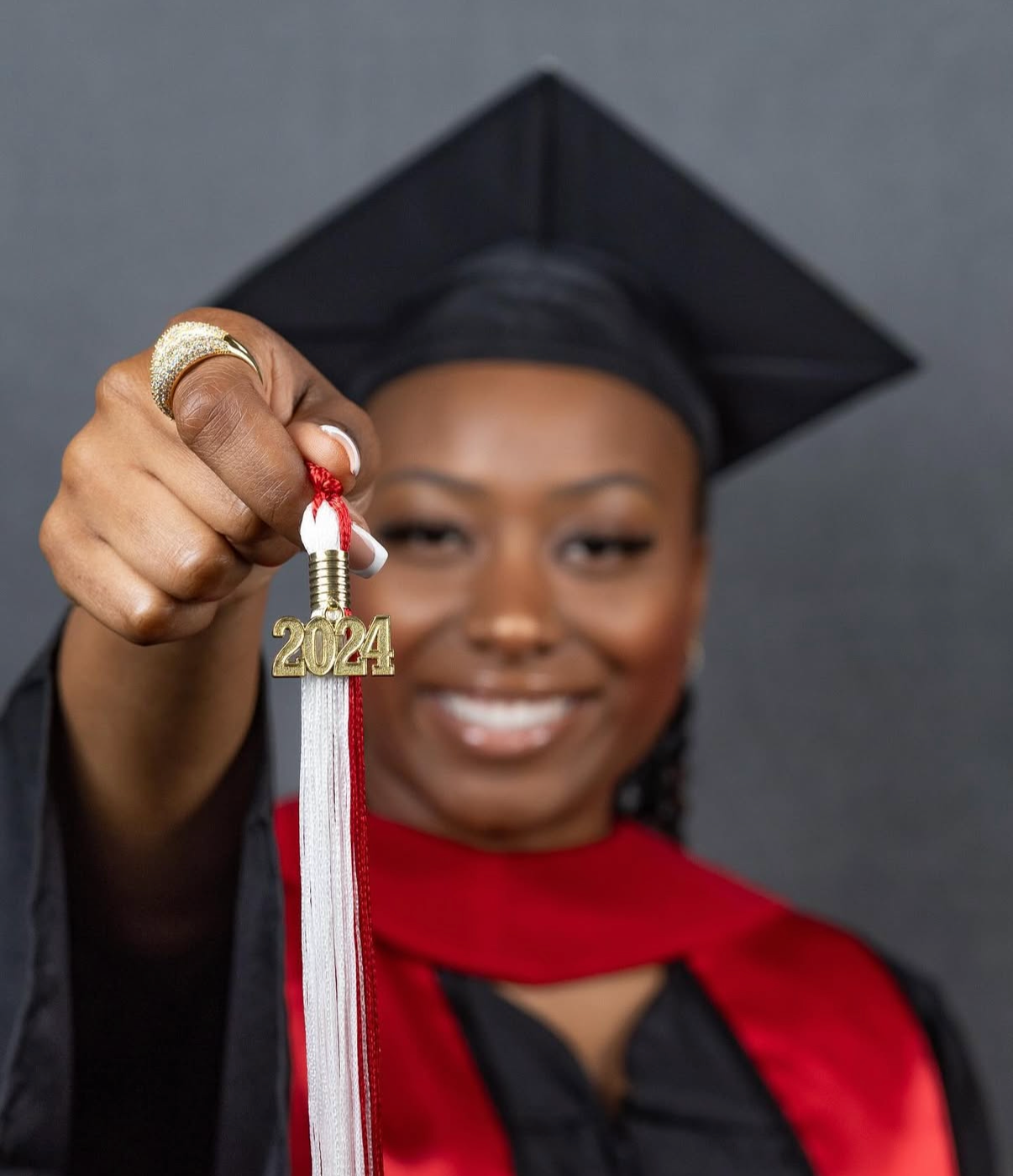 Graduate in cap and gown holds up a red and silver 2024 graduation tassel against a gray background.