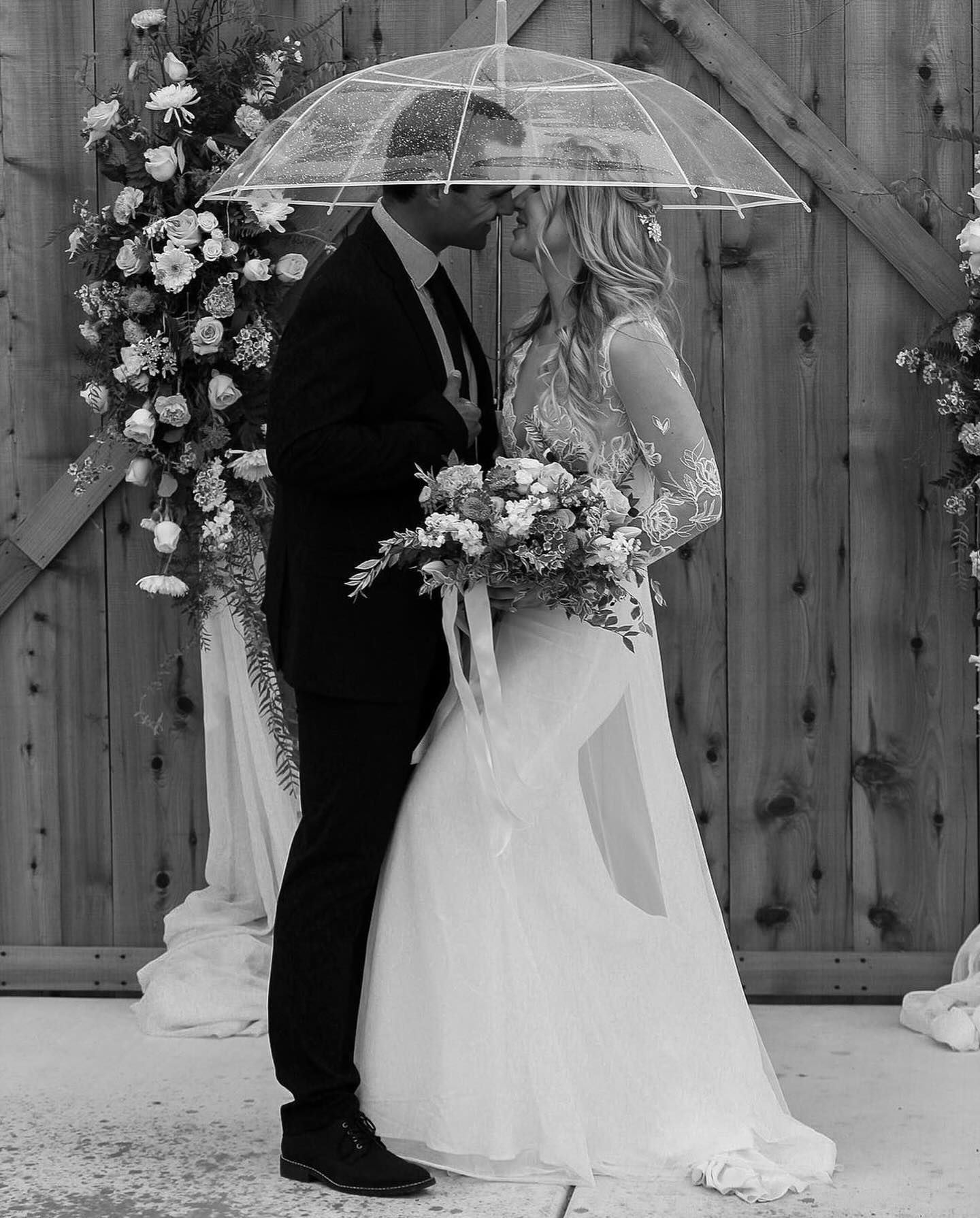 Wedding couple under clear umbrellas with floral decorations against wooden fence backdrop.