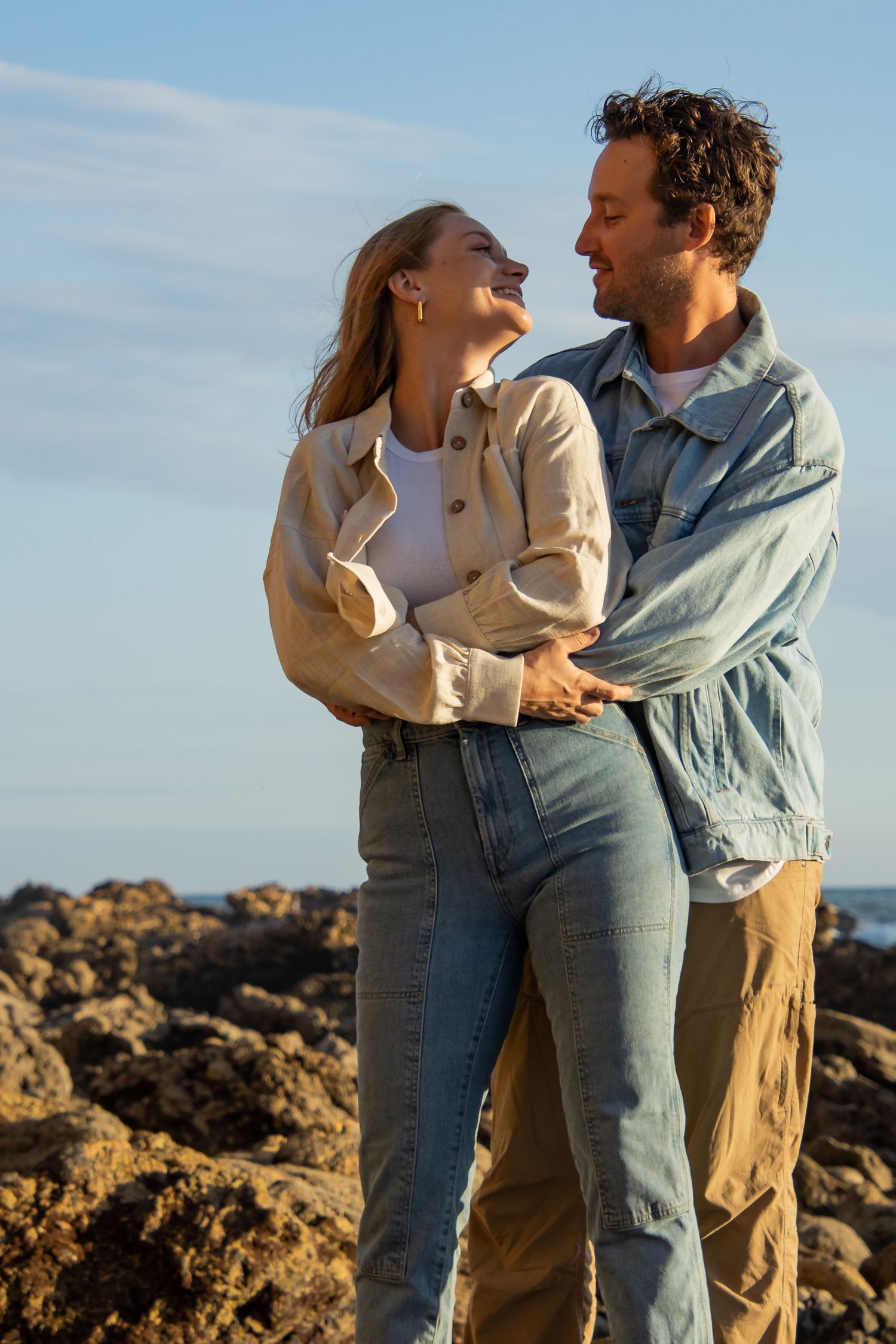 Romantic couple embracing and laughing together on rocky coastal landscape at sunset in casual denim outfits.