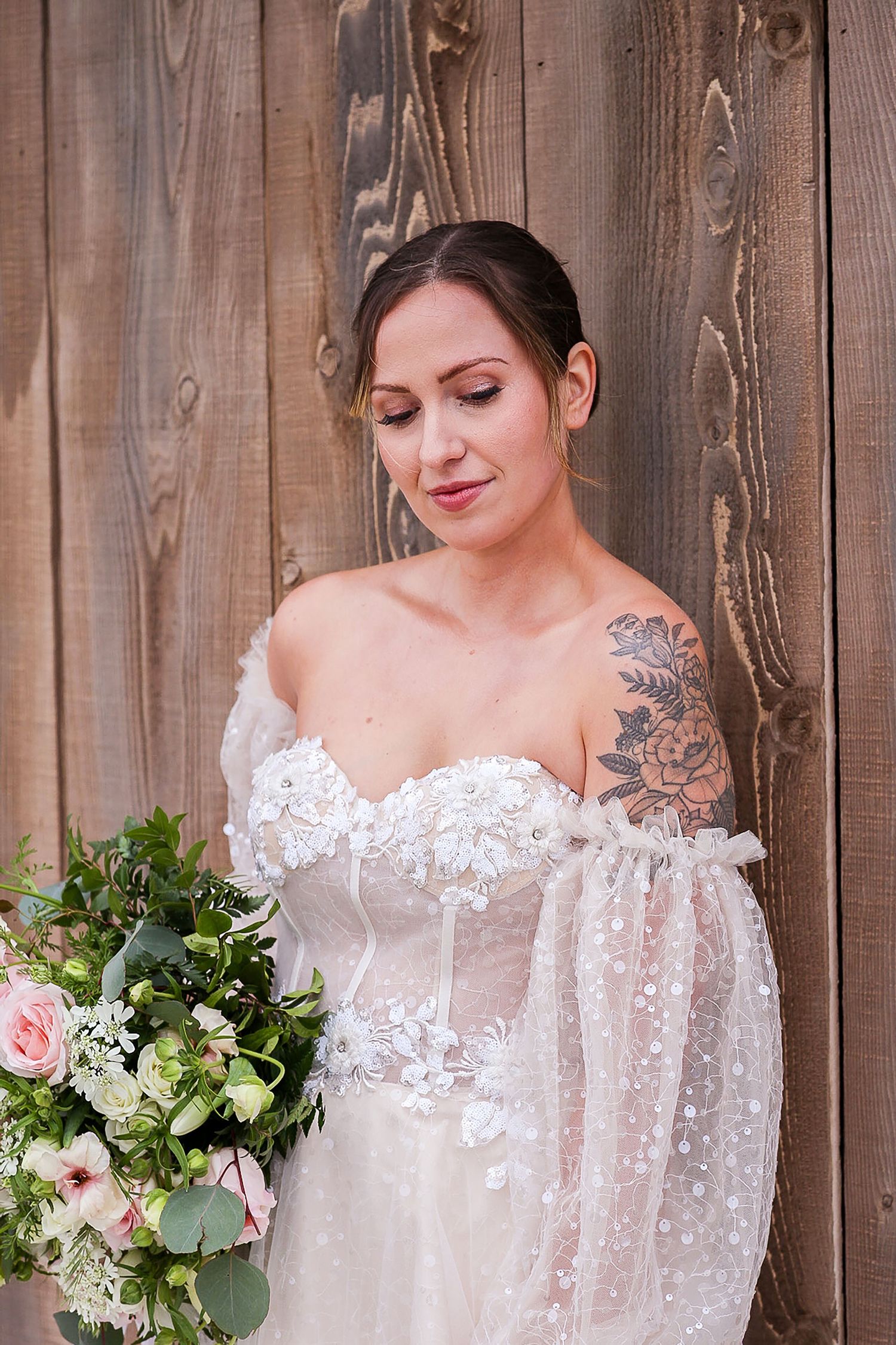 Bride in lace off-shoulder wedding dress holding bouquet against wooden wall background.