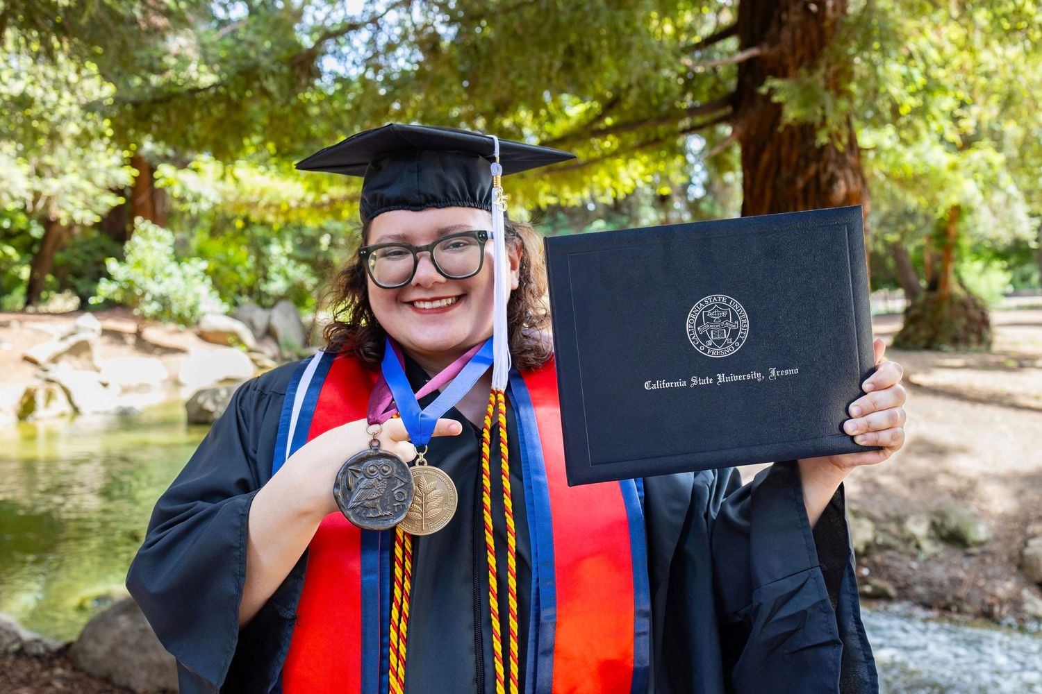 Graduate displaying diploma with academic regalia and honor cords in outdoor setting.