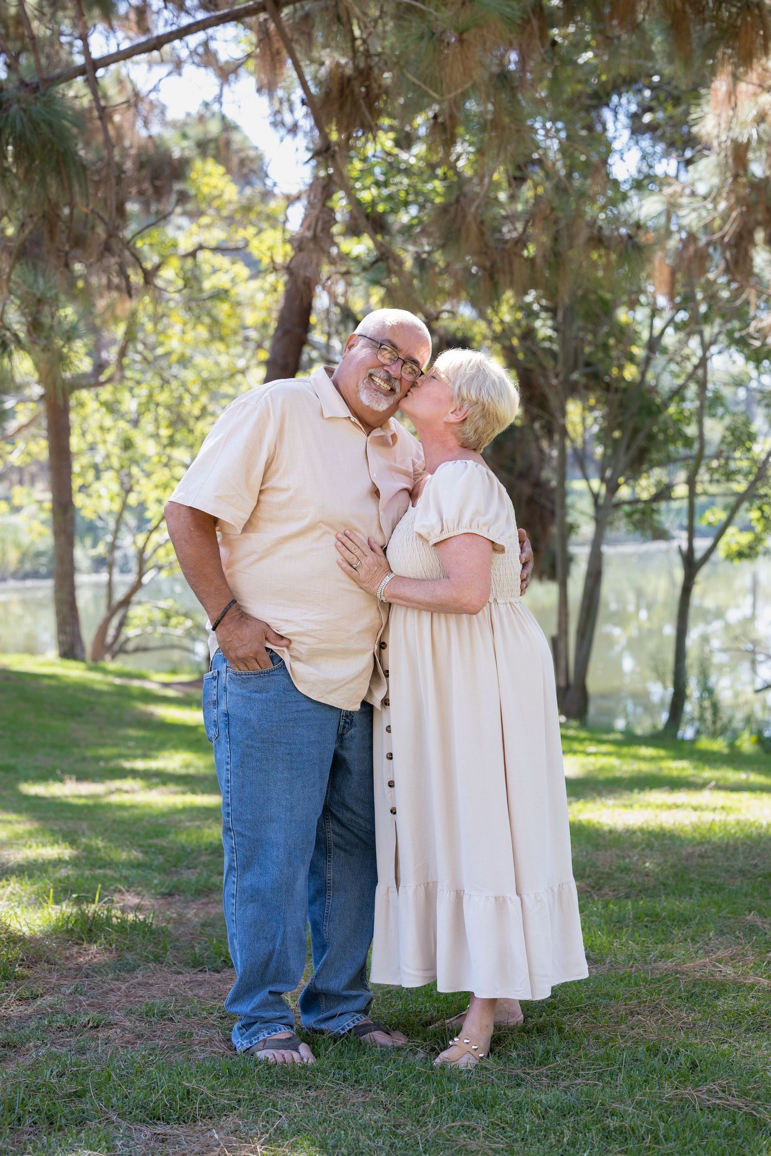 A couple shares tender moments in a park setting with trees and grass on a sunny afternoon.
