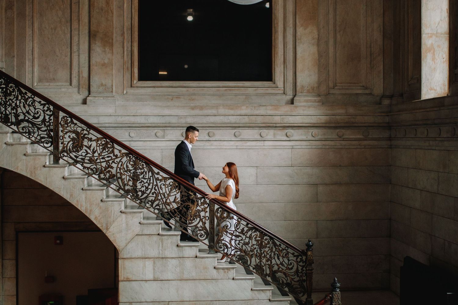 Couple poses on The Ellicott Square Building Staircase holding hands during their engagement session.