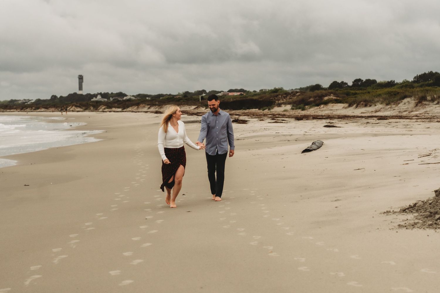 Couple holding hands walking on a sandy beach under cloudy skies with dunes and a lighthouse in the background.
