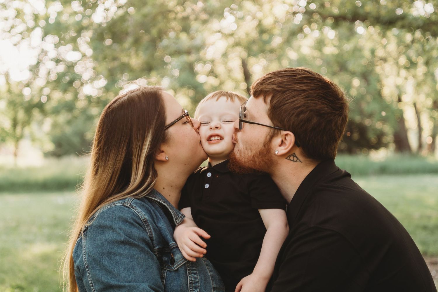 Parents kissing their laughing toddler on the cheeks outdoors in a sunny green park.