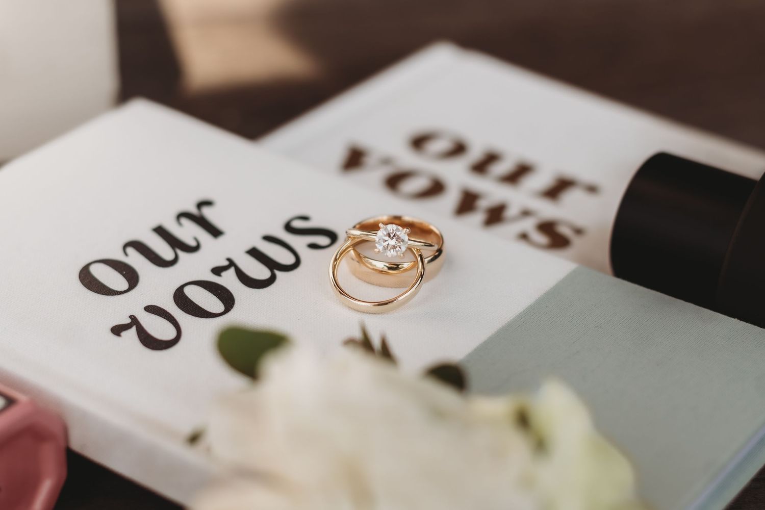Two gold wedding rings and a diamond engagement ring rest on an open vows book beside a white flower.
