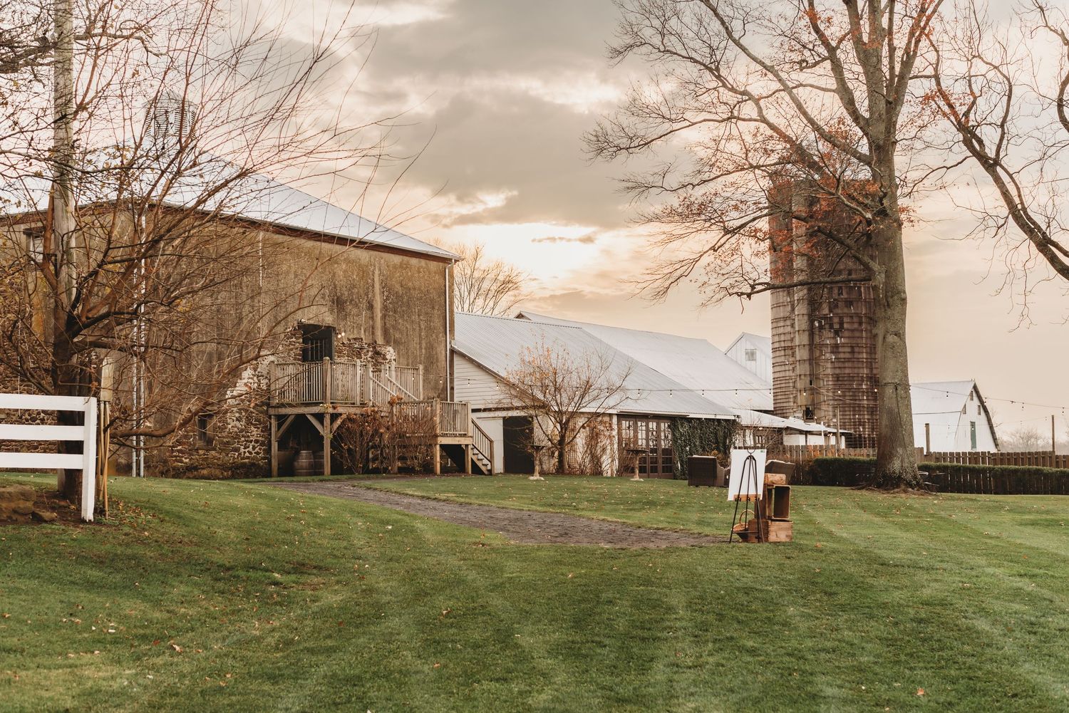 A panoramic view of a rustic barn and farmland during sunset with bare trees in the foreground.