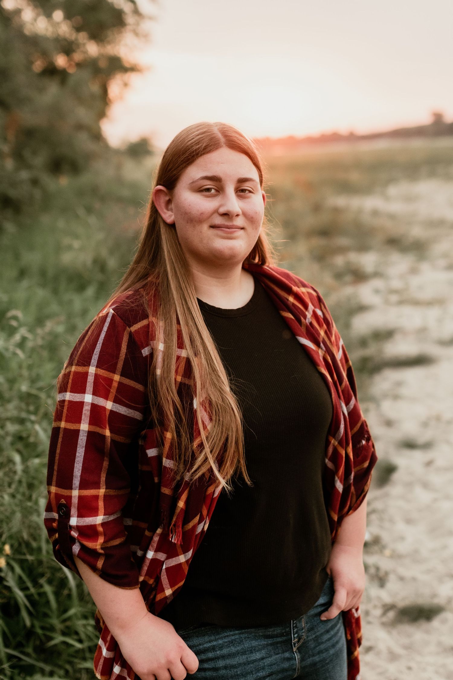 Person in plaid shirt and black top poses outdoors during golden hour.