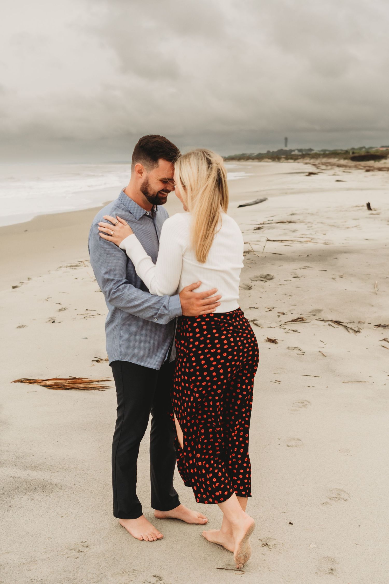 Couple embracing romantically on a sandy beach with overcast skies, woman in floral skirt and man in blue shirt.