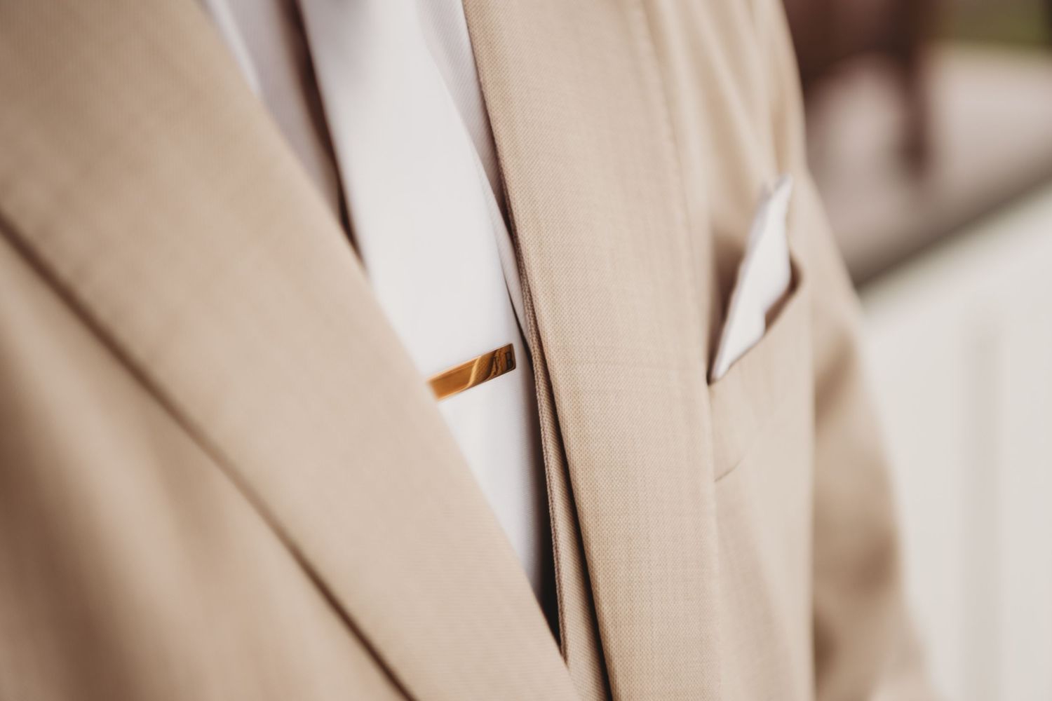 Close-up of a beige suit jacket with a gold tie clip and white pocket square, perfect for formal occasions.