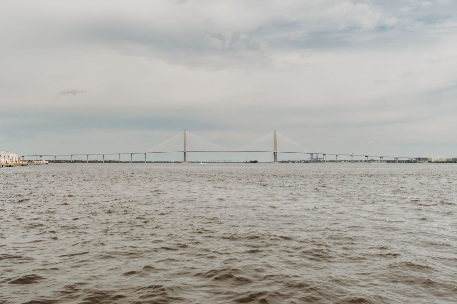 A cable-stayed bridge spans a wide river under a cloudy sky, with choppy waters in the foreground.