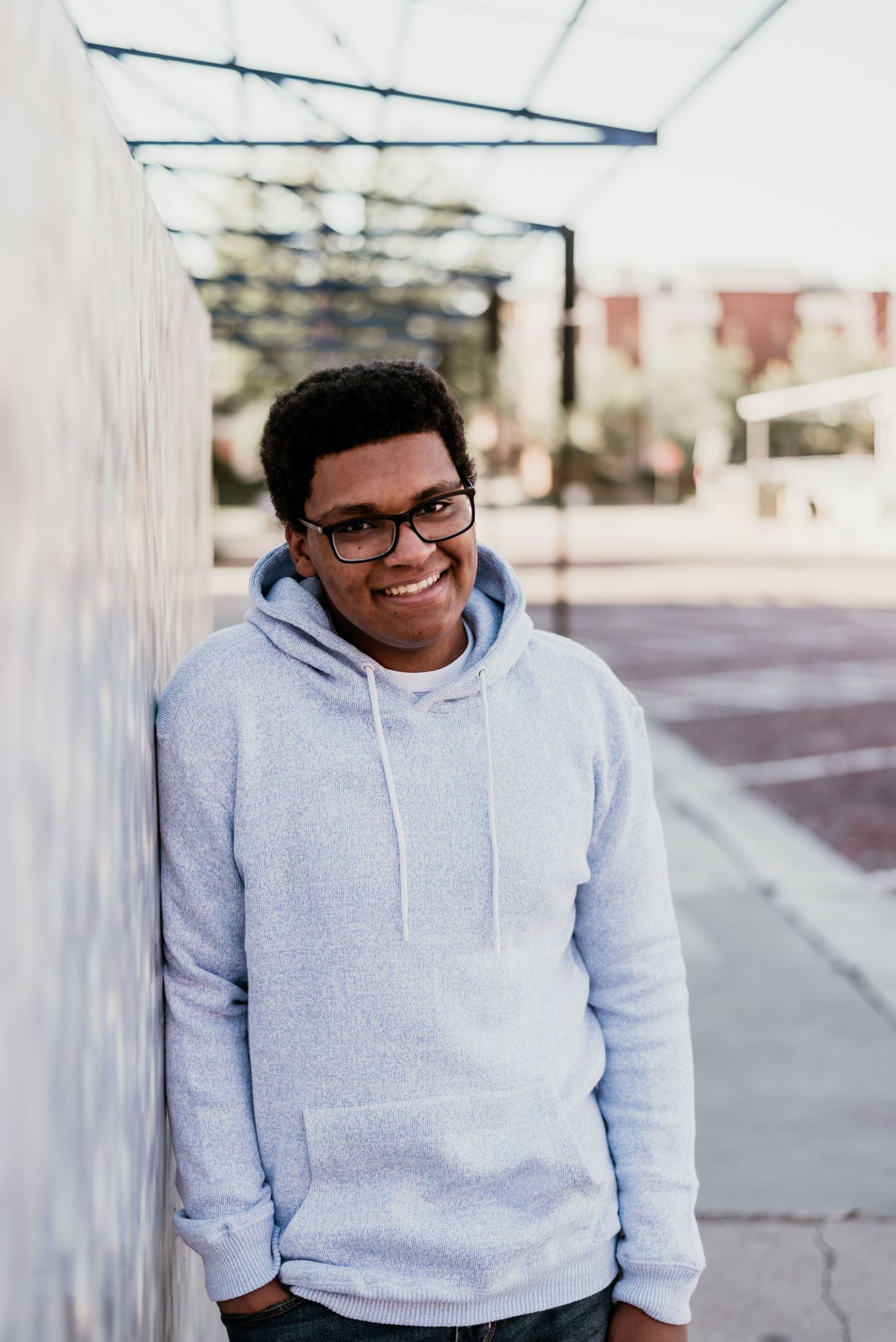 A person in a light gray hoodie and glasses sits against a white wall outdoors.