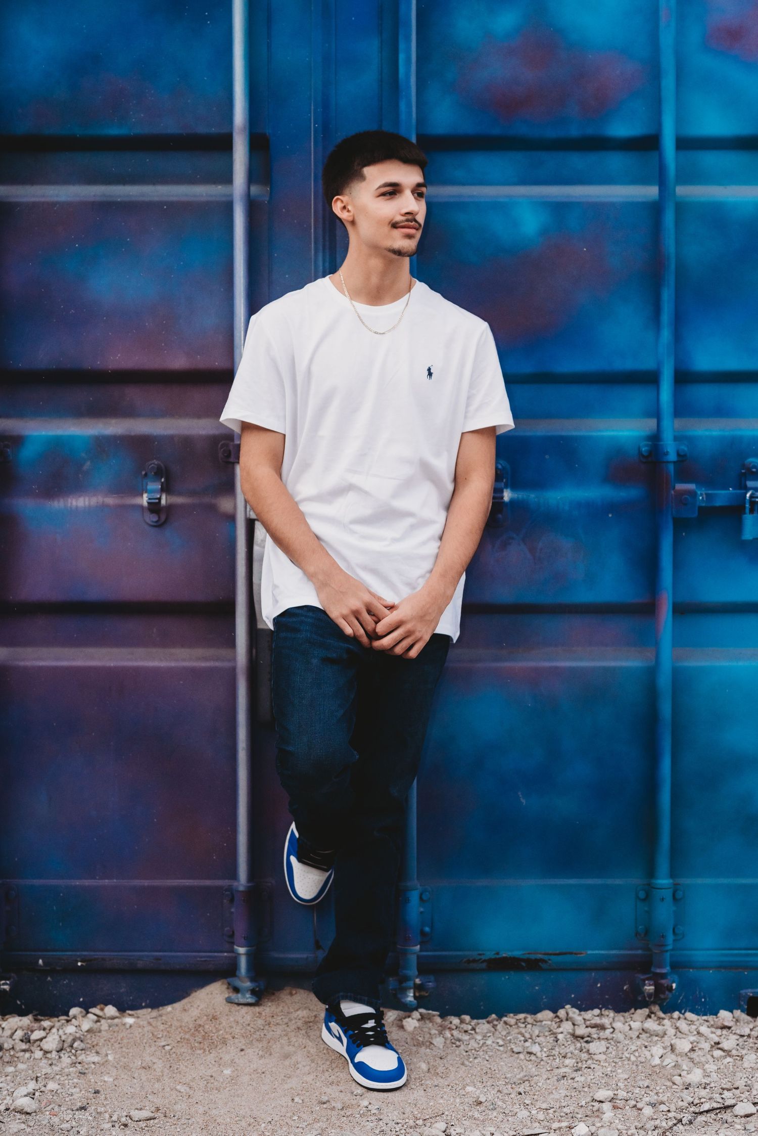 A person in white t-shirt sits against a blue industrial backdrop with gravel ground.