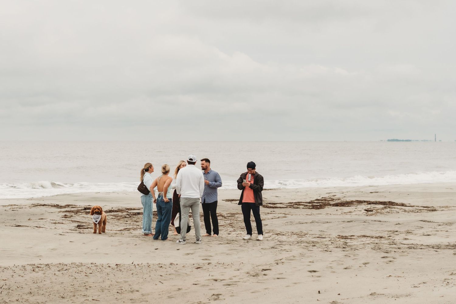 Group of friends and a dog gathered on a sandy beach under an overcast sky near the ocean shoreline.